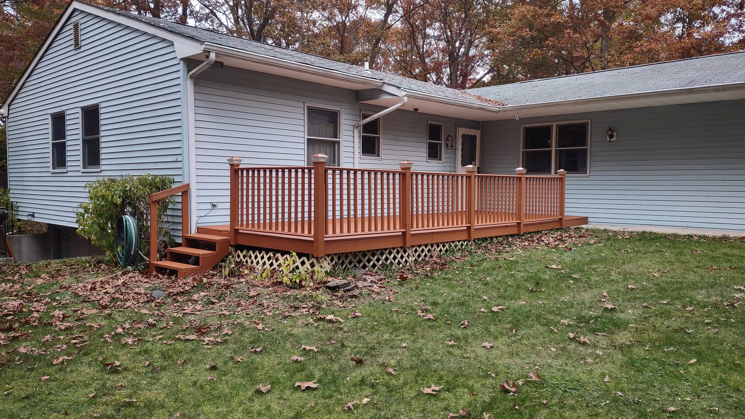 Backyard view of a light blue house with a newly built wooden deck with railing and stairs, green grass, fallen leaves, and trees with autumn foliage in the background.