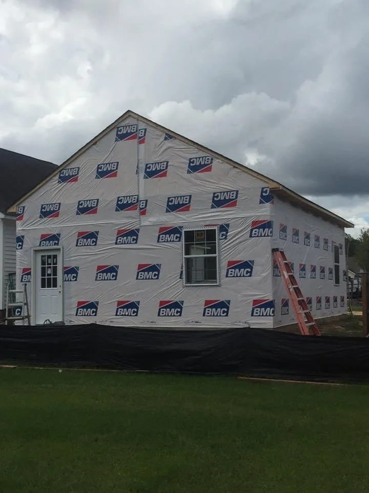 A house under construction with BMC house wrap on the exterior walls, a door, two windows, and a ladder leaning against the wall. Overcast sky in the background.