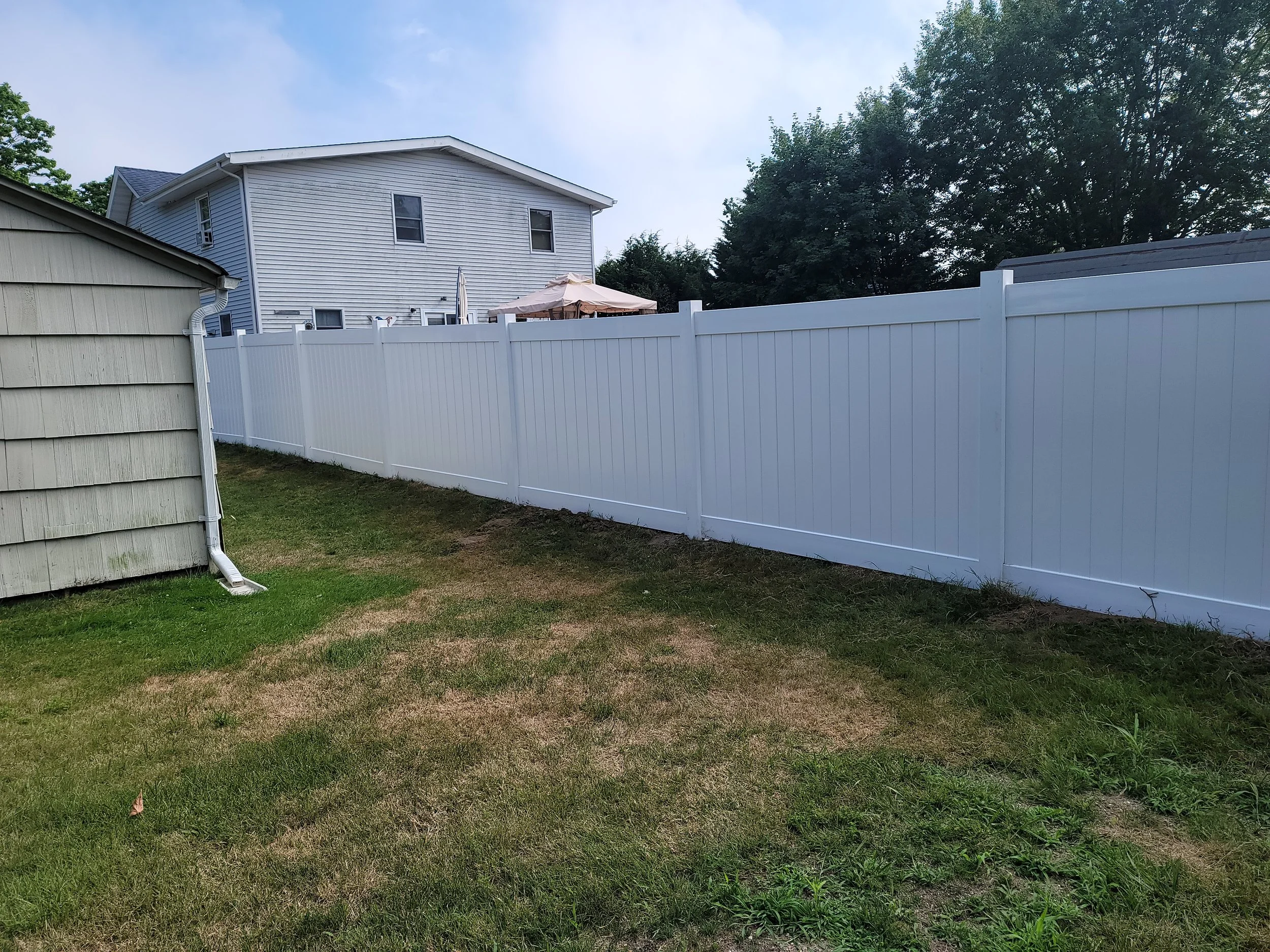 Backyard with a white vinyl fence, a small patch of grass, and a beige shed on the left. In the background, there is a blue multi-story house with trees behind it.