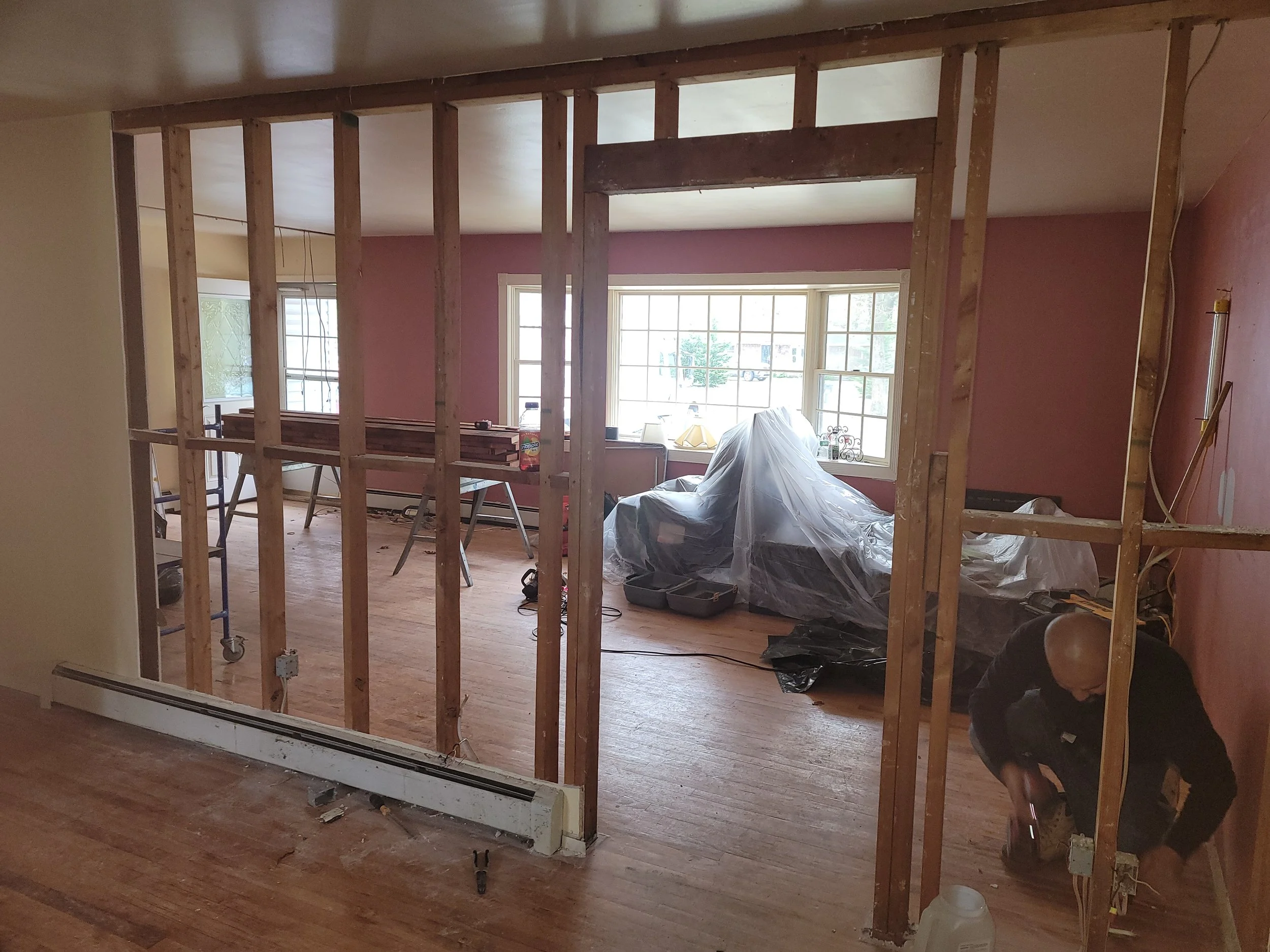 Living room under renovation with exposed wooden framing, a man working on electrical wiring, covered furniture, and a large bay window letting in natural light.