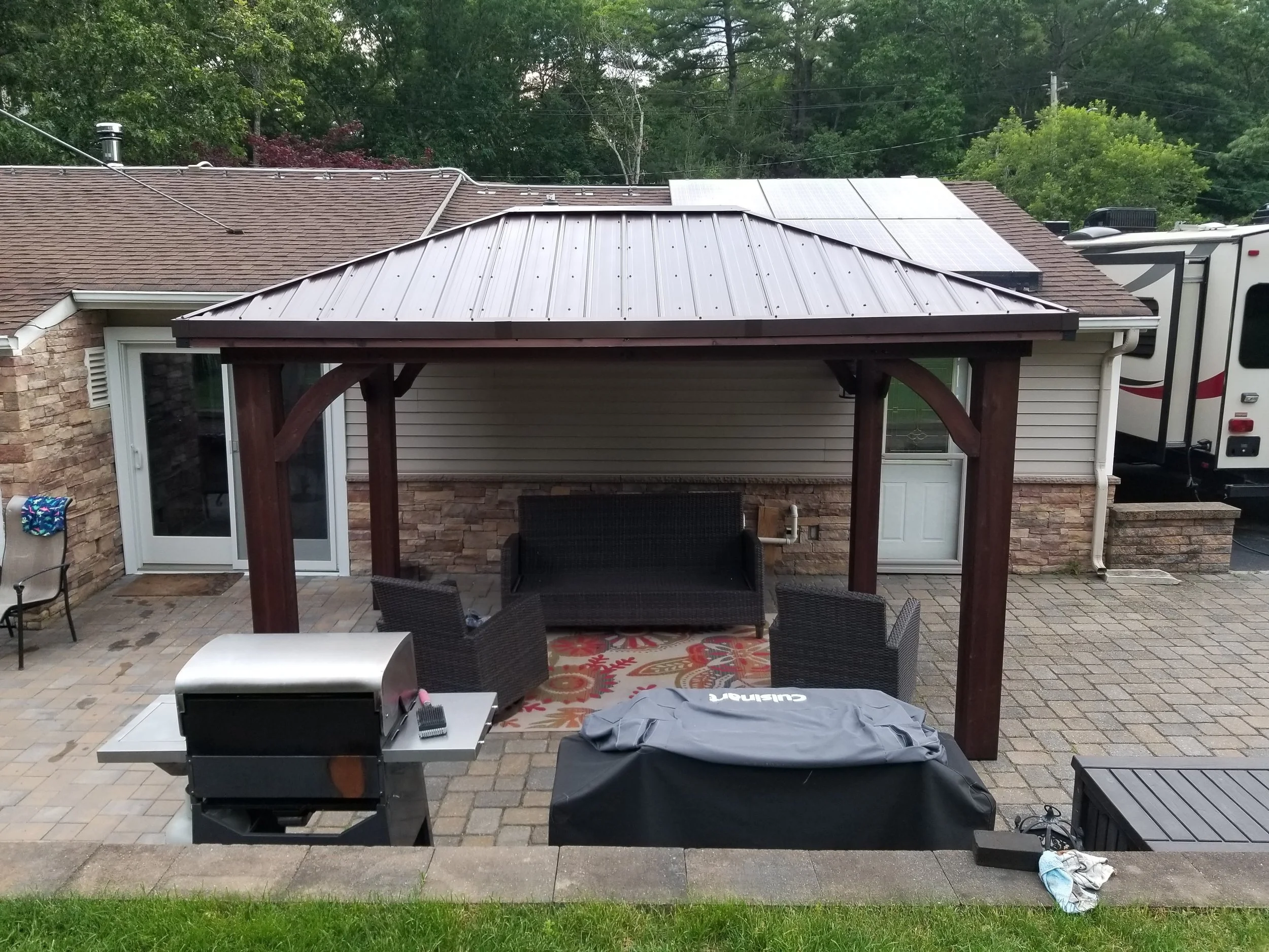 Backyard patio with a covered pergola, outdoor seating, a grill, and a patterned rug, with solar panels on the roof of the house in the background.
