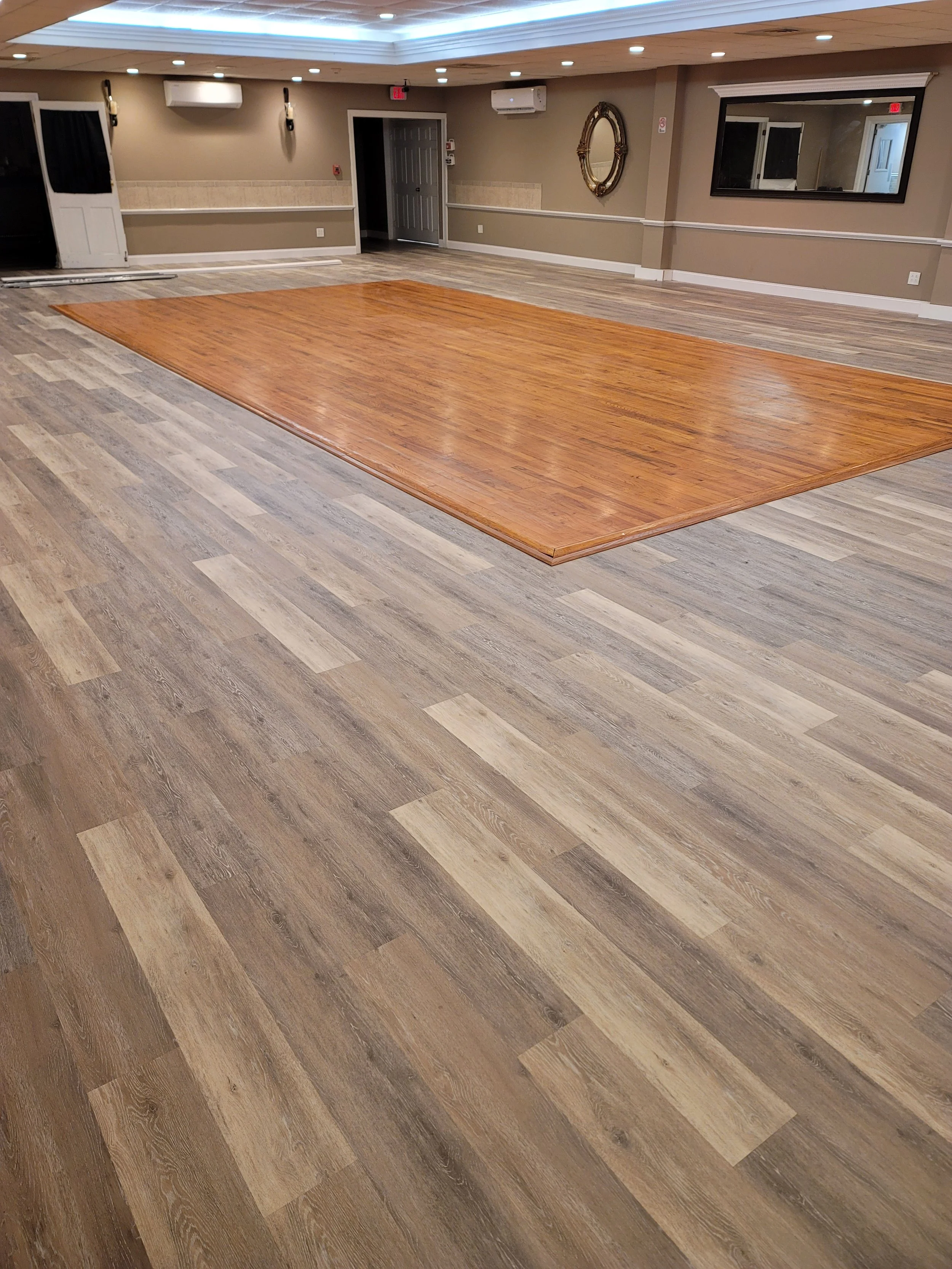 Empty indoor space with wood and laminate floors, beige walls, a large mirror, and a wooden dance floor area.