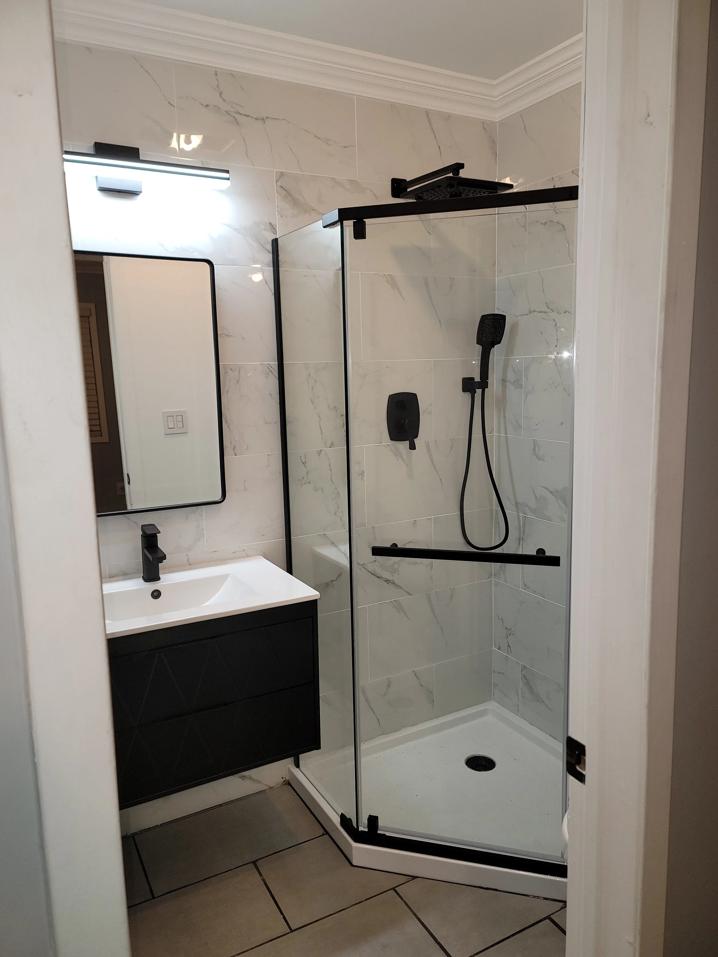 Modern bathroom with a glass-enclosed shower, black fixtures, a white sink with a black vanity, a large mirror, and beige marble-like tiled walls.