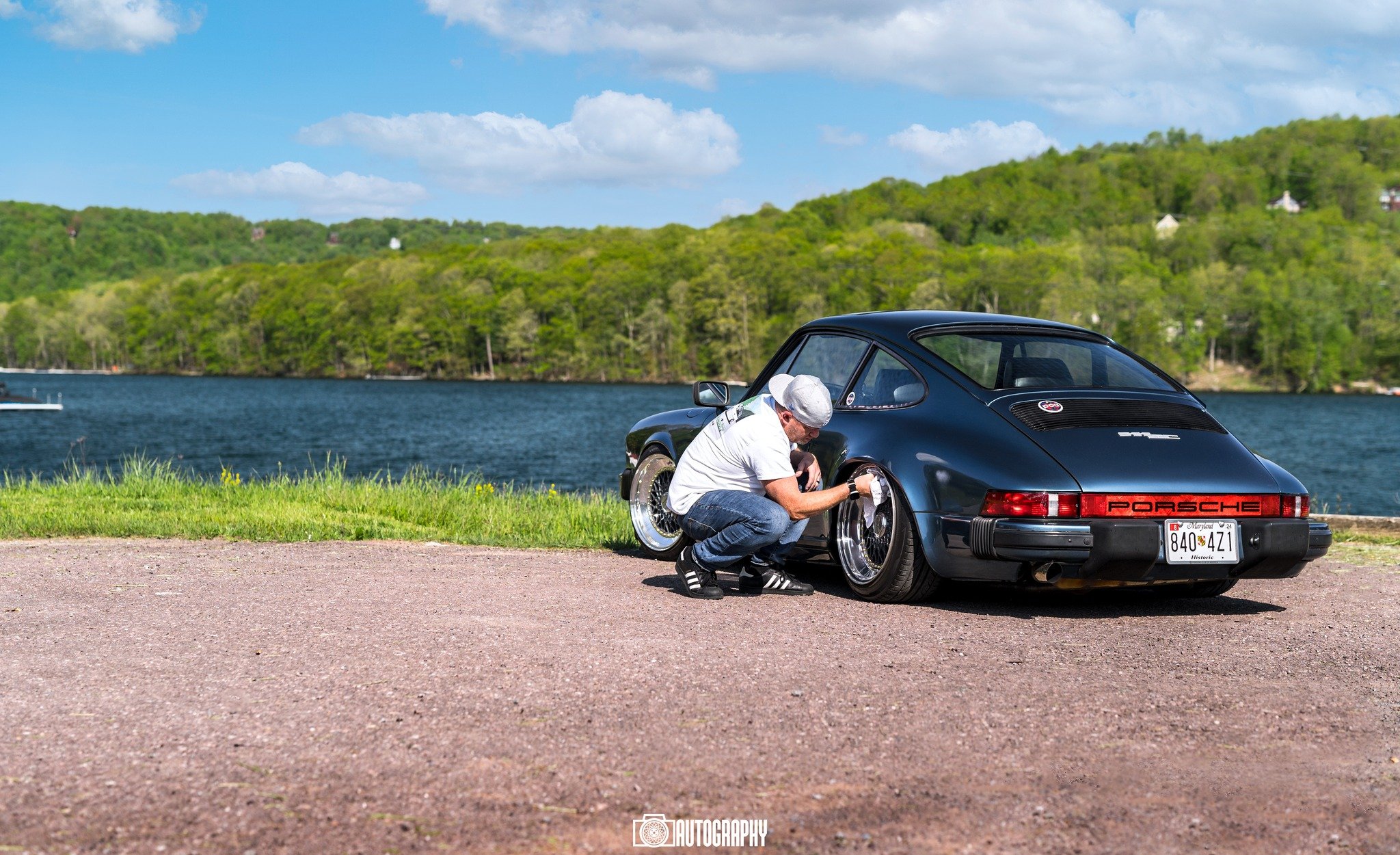 A man fixing a black Porsche 911 parked near a lake on a sunny day with green trees and hills in the background