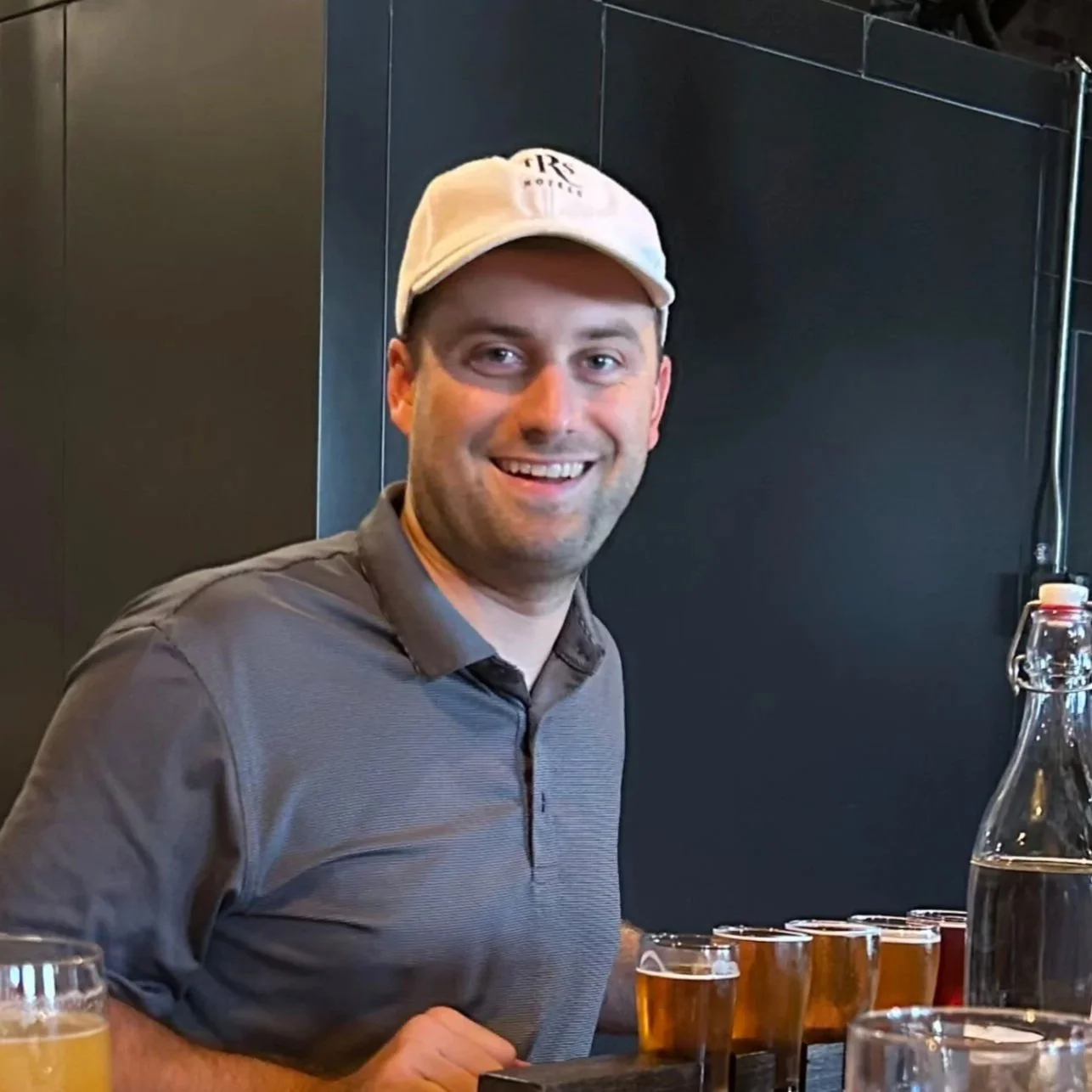 A smiling man wearing a white cap and a gray polo shirt sitting at a bar with glasses of beer and a water bottle in front of him.