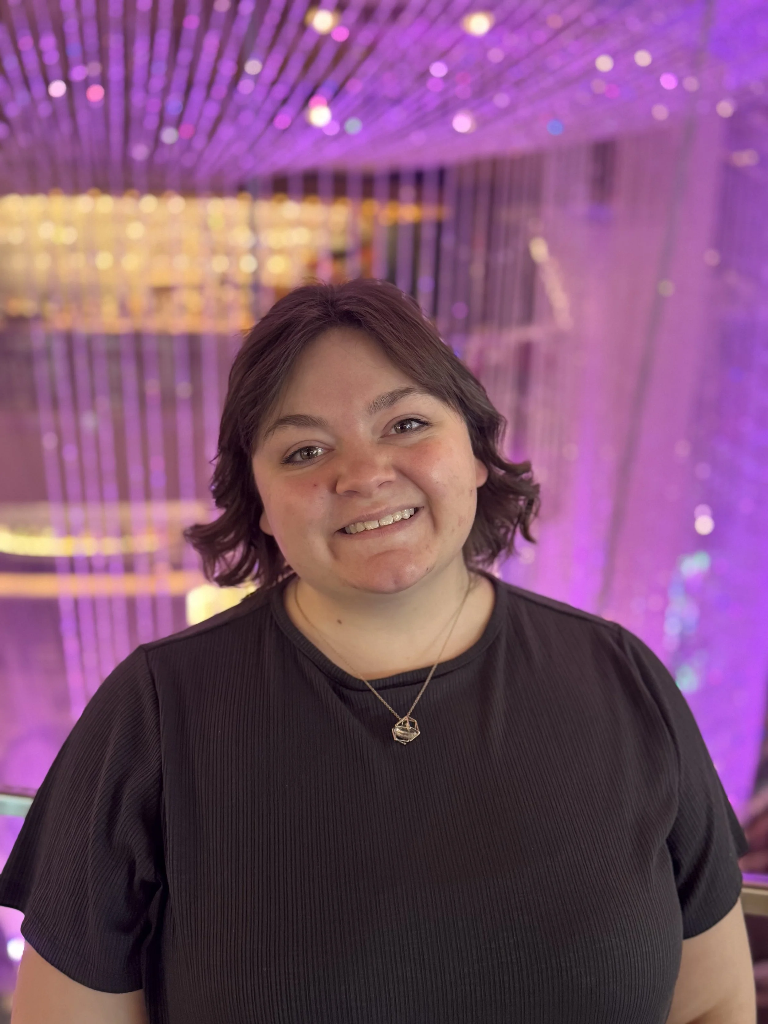 A smiling woman with short dark hair standing in front of purple and gold shimmering lights.