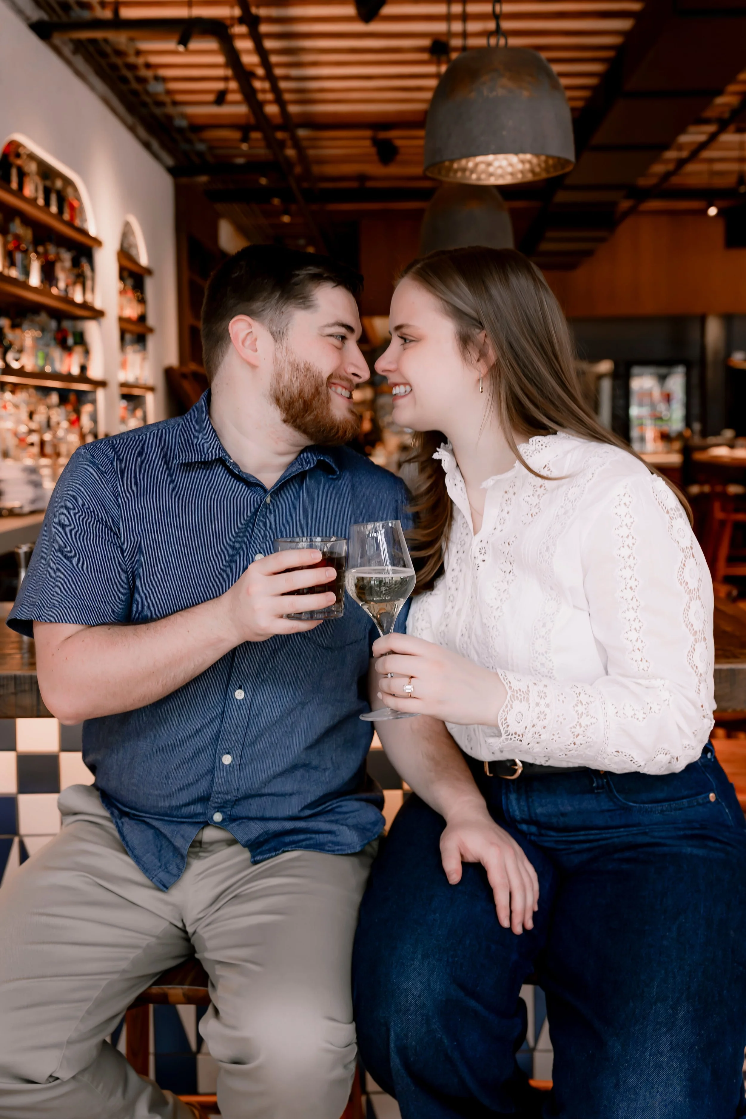 A couple sitting closely together, smiling and touching noses, holding glasses of wine, in a cozy bar or restaurant setting.