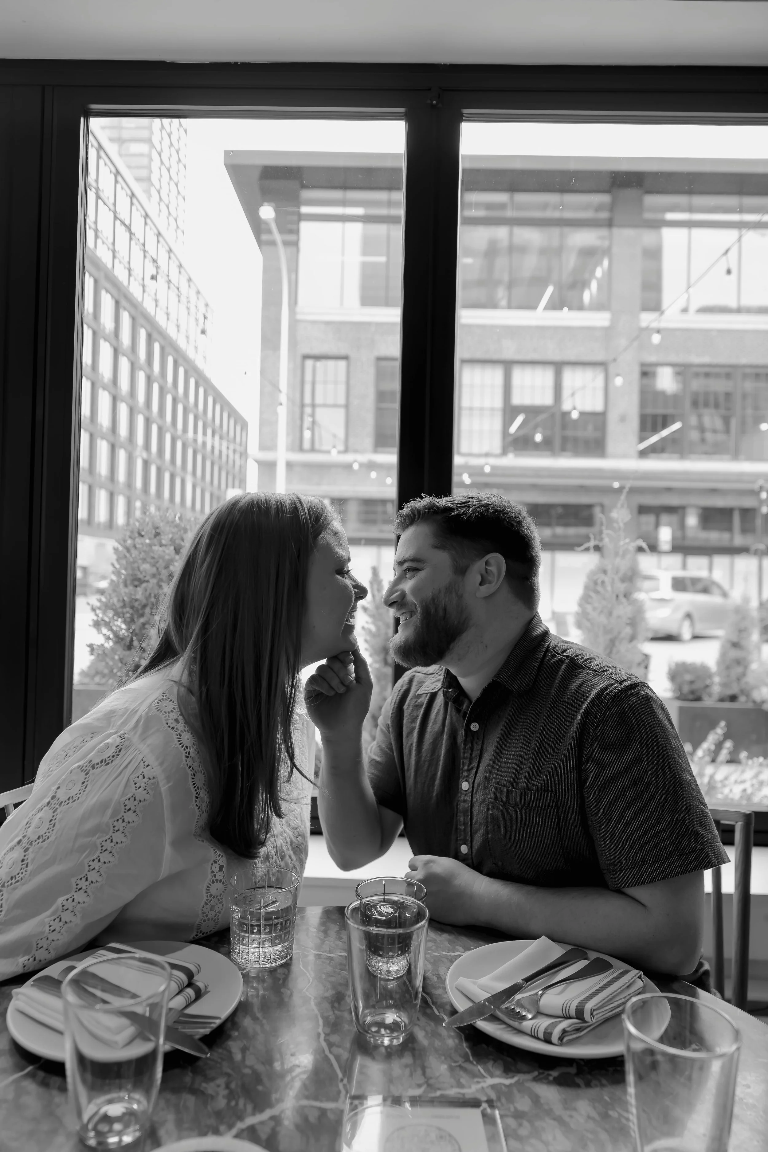 A black-and-white photo of a couple sitting at a restaurant table, smiling and leaning close to each other. The table is set with glasses, plates, napkins, and cutlery. Large windows behind them reveal a cityscape with buildings, street lamps, and tr