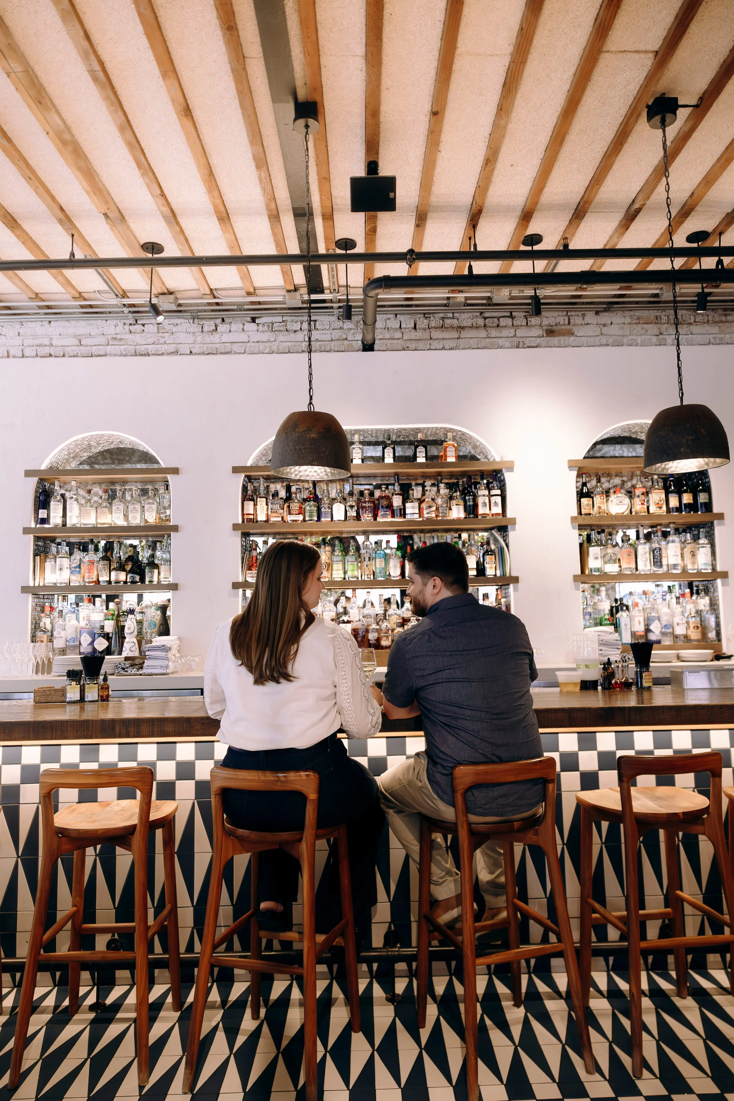 A couple sitting at a bar counter in a modern restaurant or bar, with various bottles of alcohol displayed behind them on shelves. The bar has a checkered tile pattern on the front, and there are large pendant lights hanging from a wood-beamed ceiling. The couple appears to be engaged in conversation, and the overall ambiance is warm and stylish.