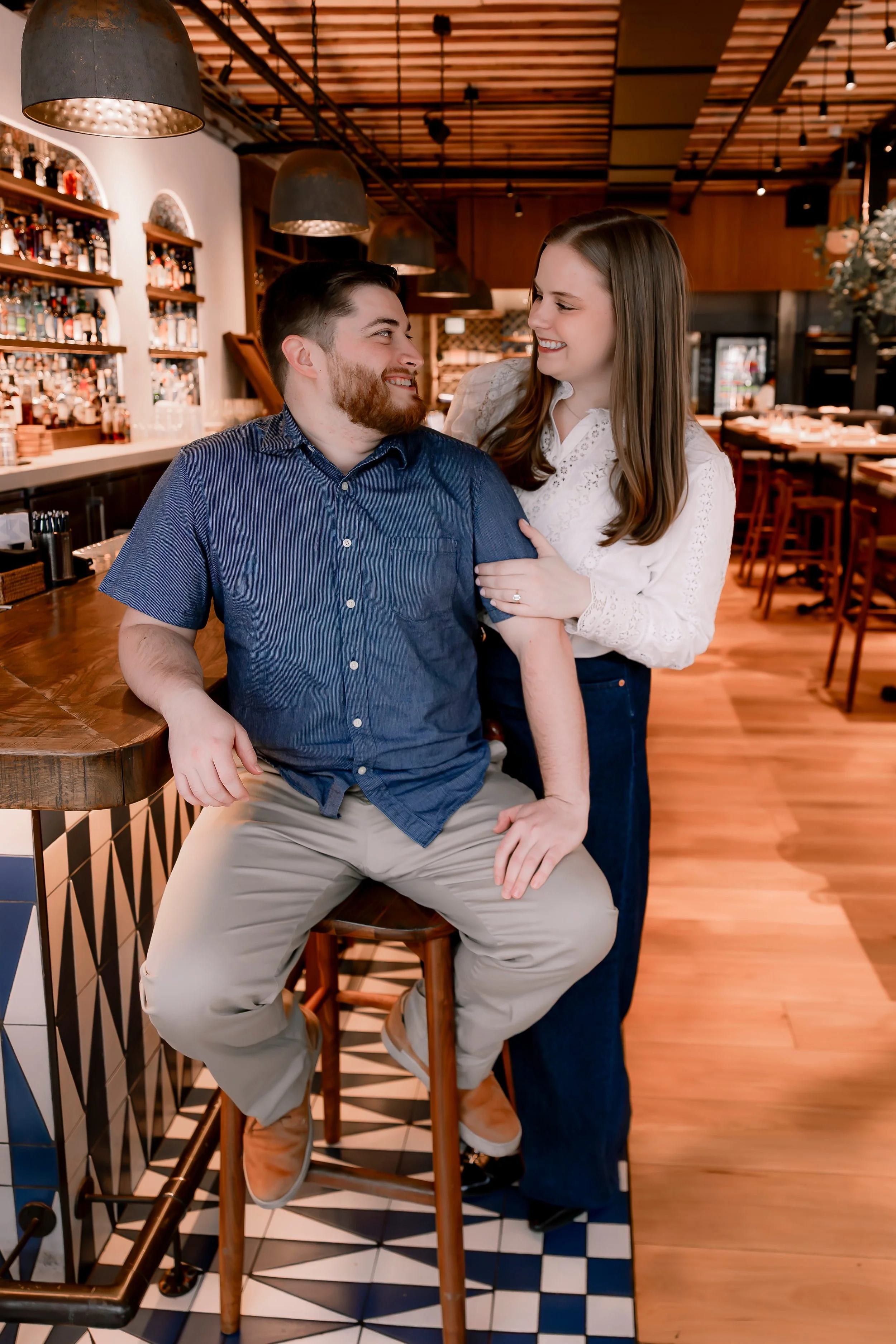 A couple is smiling and looking at each other in a cozy bar or restaurant with wooden decor, shelves full of bottles, and warm lighting.