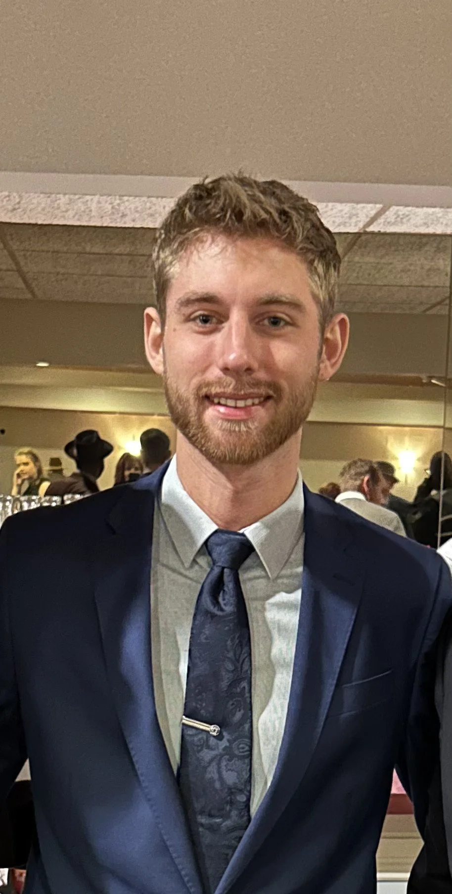 A young man with light skin, short curly hair, and a beard, wearing a navy blue suit, light shirt, and patterned tie, smiling at a social event with people in the background.