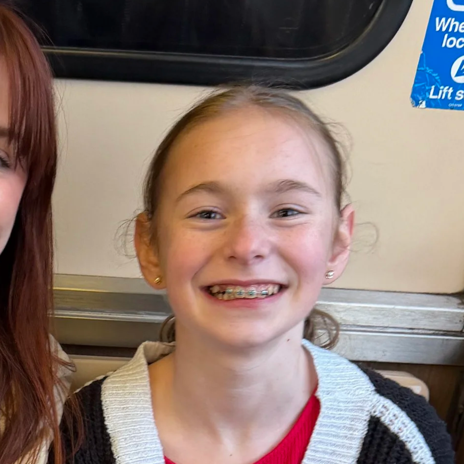 A young girl with braces smiling, wearing a red shirt and a black and white sweater, sitting inside a bus or train.
