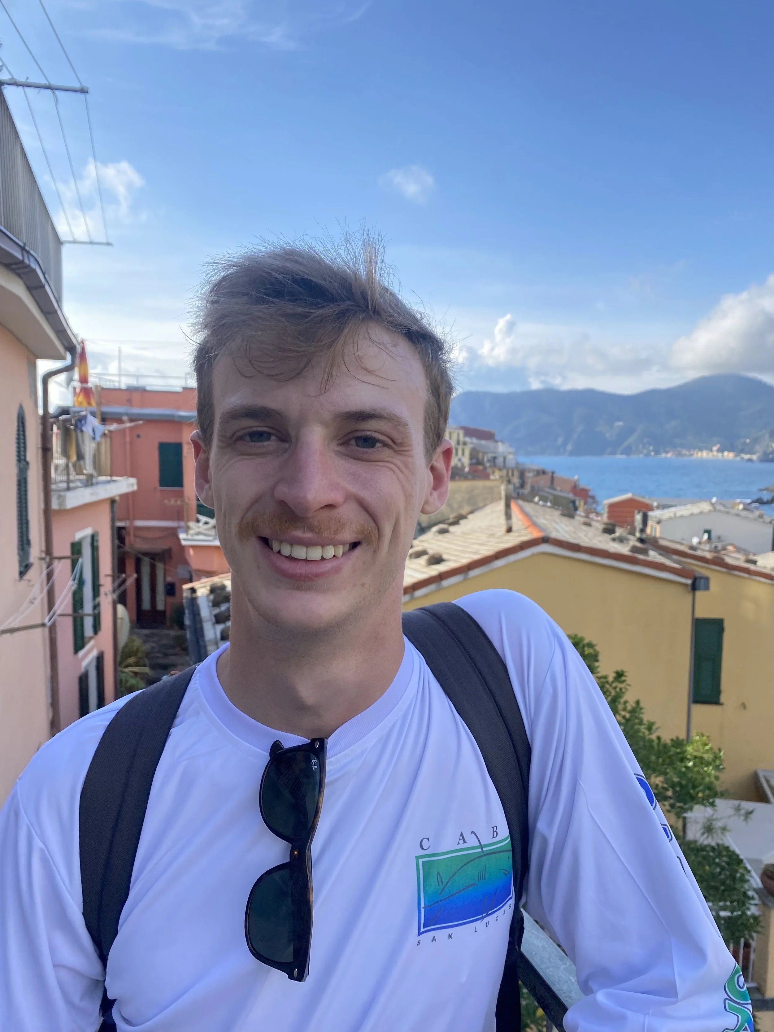 Smiling young man with brown hair standing outdoors in a coastal town with colorful buildings, mountains, and water in the background