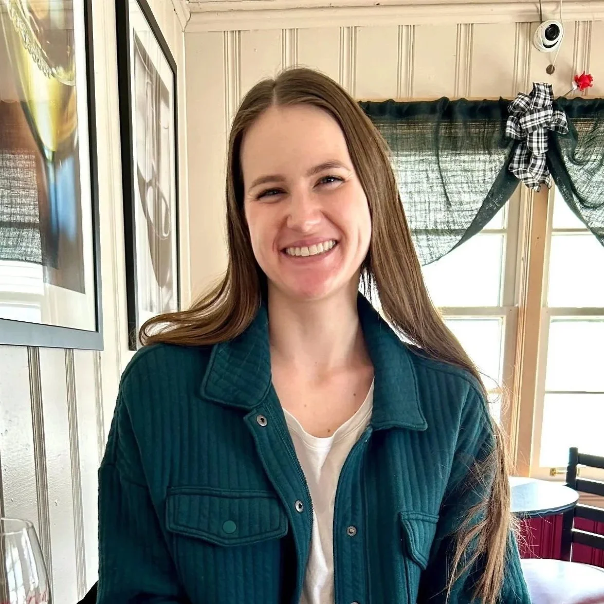 A smiling woman with long brown hair wearing a teal jacket indoors near windows with black checkered curtains tied with a checkered bow.
