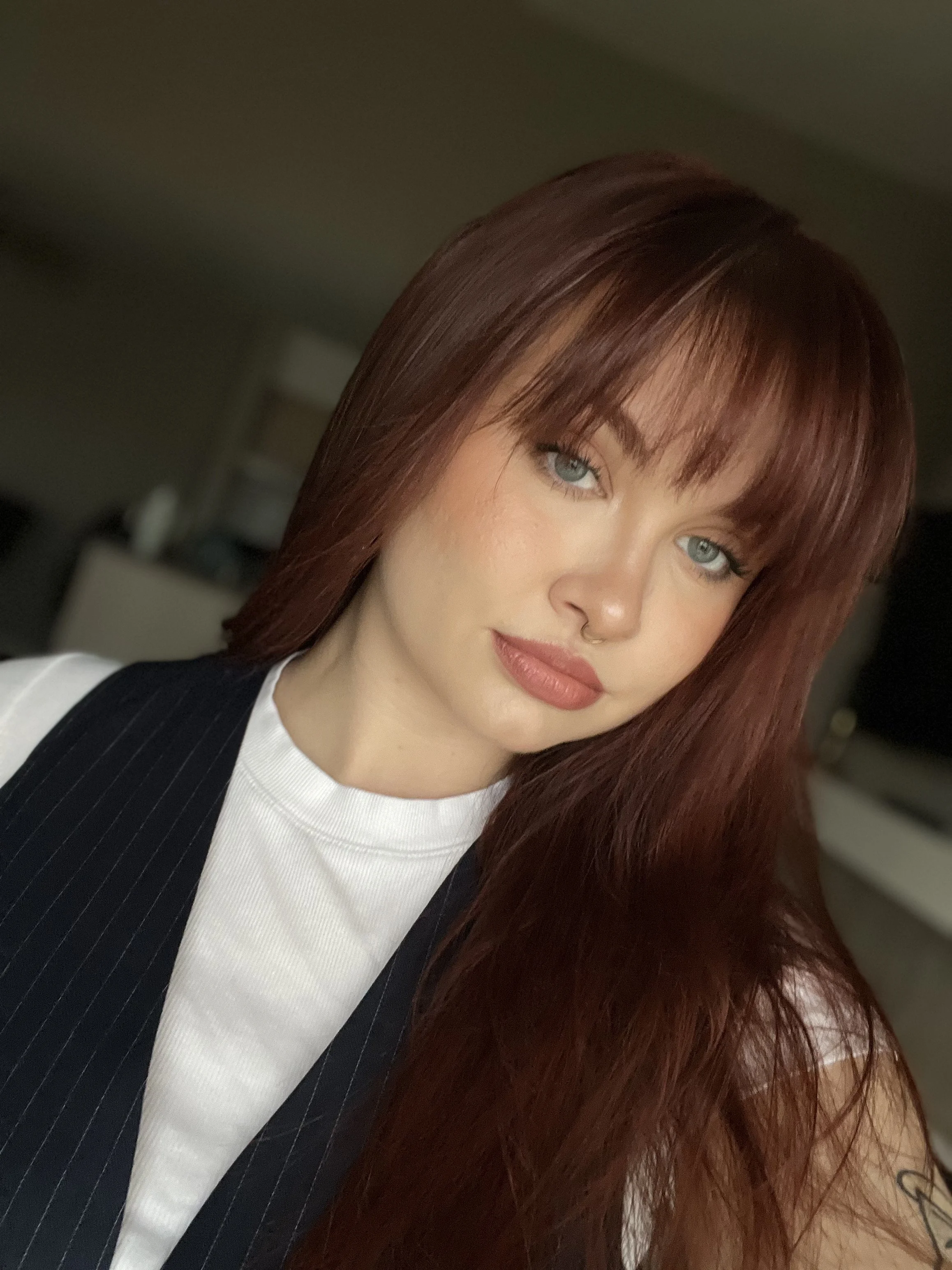 A woman with long, straight, red hair and blue eyes, wearing a white top and a black pinstripe vest, posing indoors.