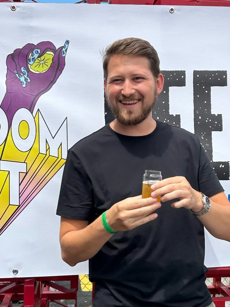 A smiling man with a beard and short hair holding a drink in a small glass at a booth with a Comic-Con themed banner in the background.