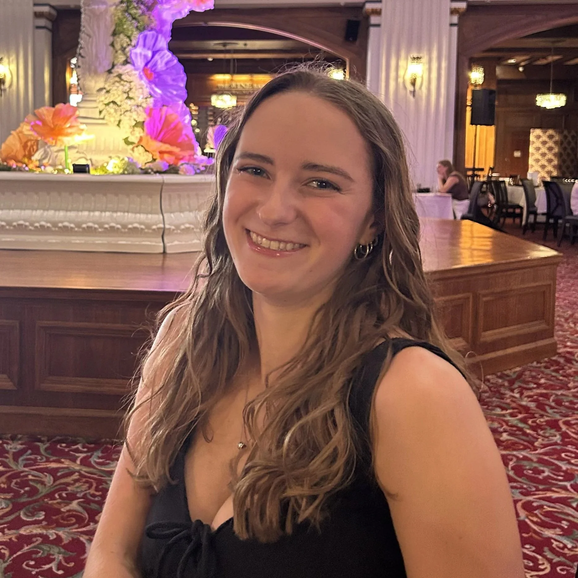 Smiling woman taking a selfie in a decorated venue with floral arrangements on a white ornate table behind her, and a wooden-paneled ceiling with chandeliers.