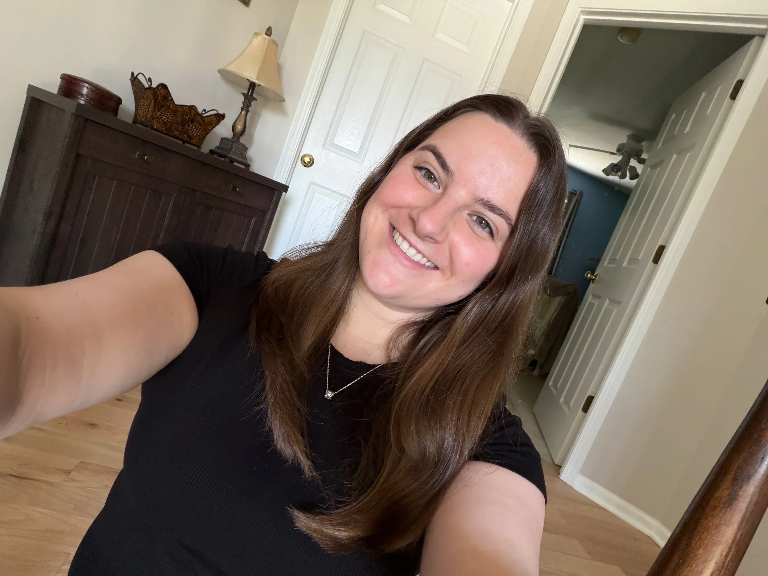 A woman taking a selfie in a room with a dark wooden dresser, a table lamp, a decorative basket, and a door open to another room with a ceiling fan.