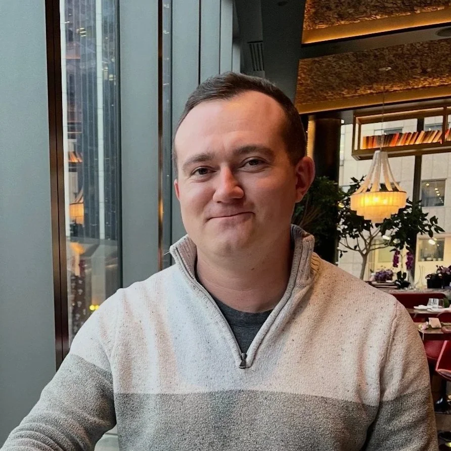 A young man with short dark hair sitting in a restaurant near large windows, smiling slightly, wearing a beige quarter-zip sweater.