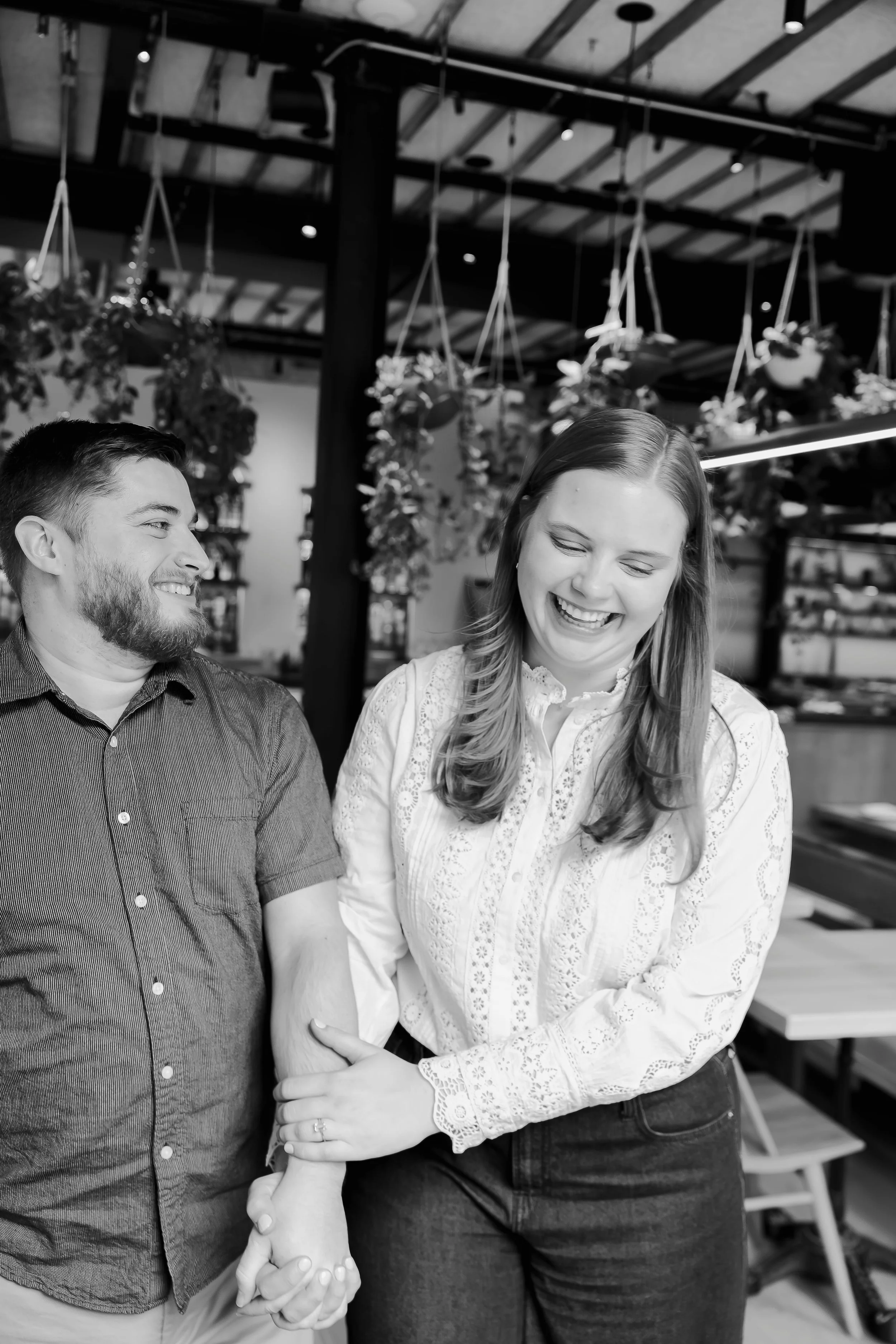 Two people, a man and a woman, smiling and holding hands in what appears to be a cafe or restaurant. The woman is laughing with eyes closed, and the man is looking at her with a smile. Hanging plants are visible in the background.