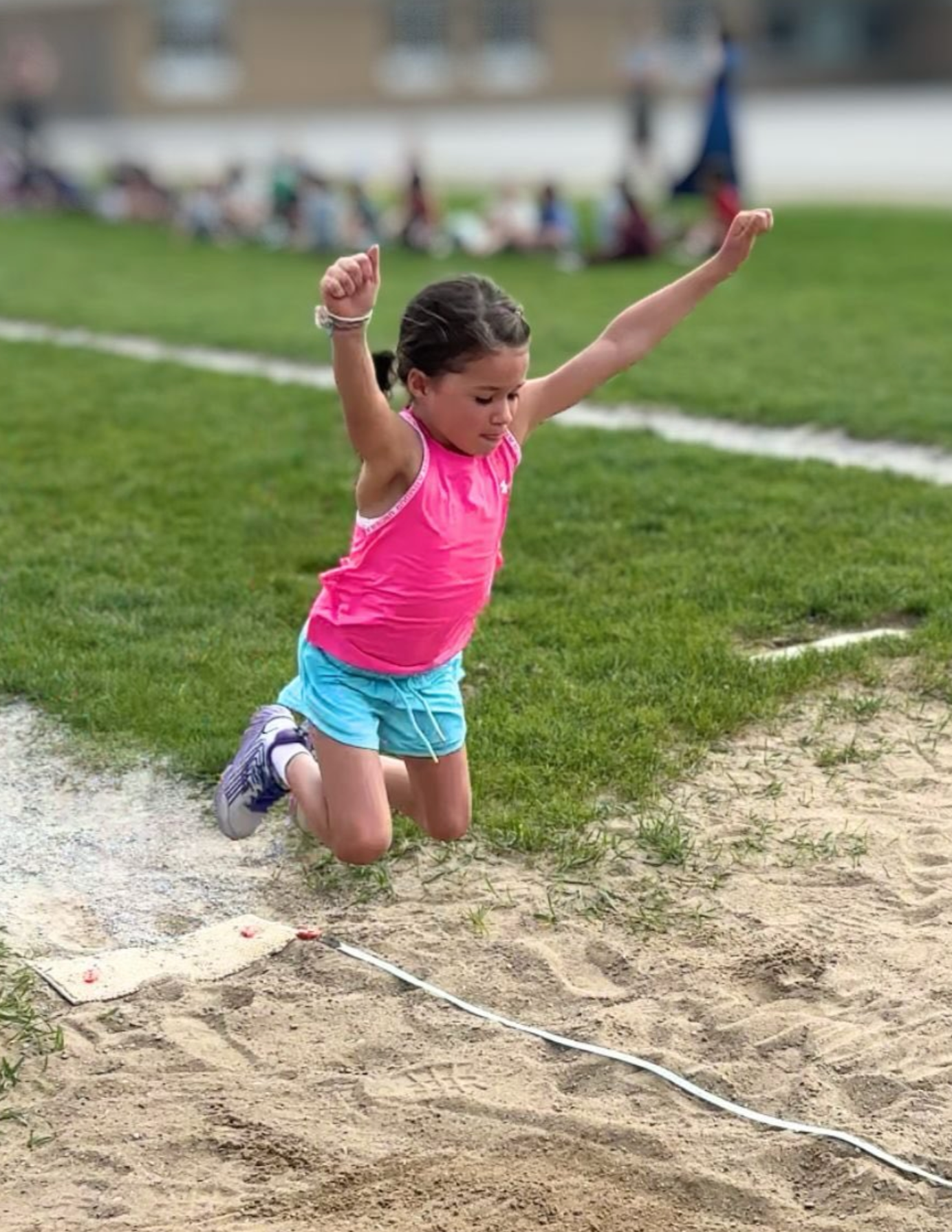 A young girl in a pink tank top and blue shorts is jumping into a sandpit during a track and field event, with a blurred crowd in the background.