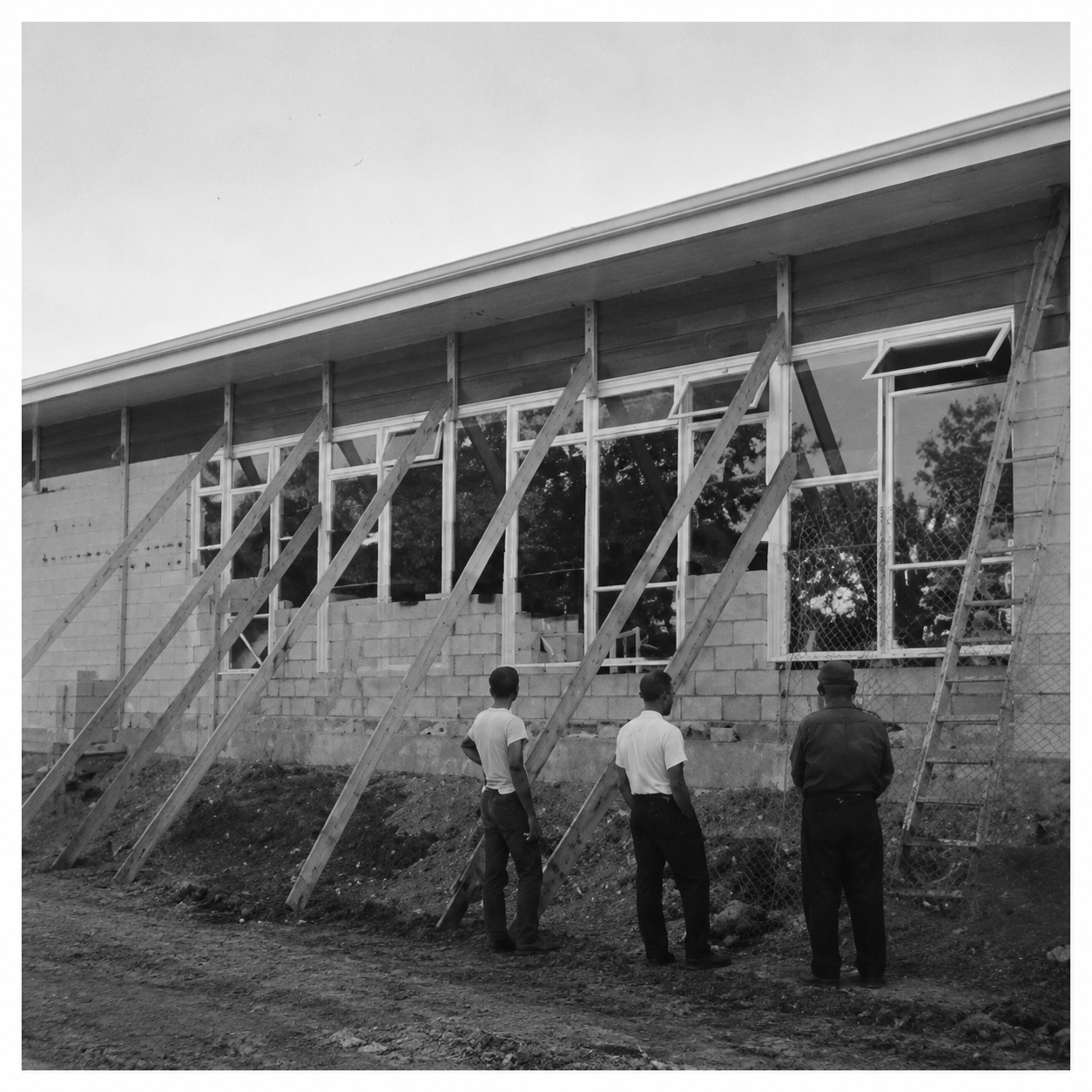 Three men stand outside a building under construction, observing wooden supports and scaffolding along the wall.