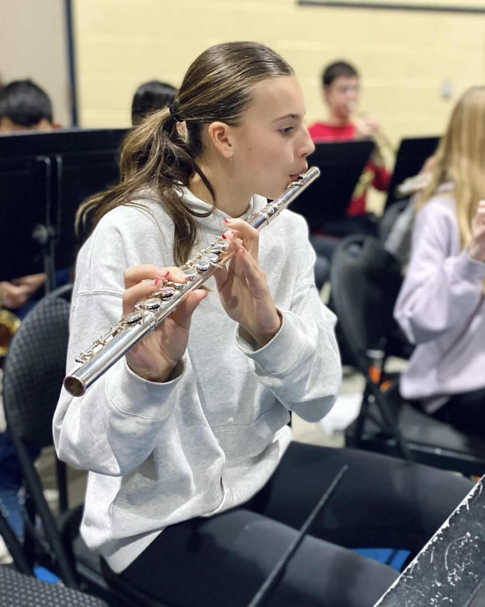 A teenage girl in a gray hoodie playing a silver flute in a classroom setting, with other students in the background.