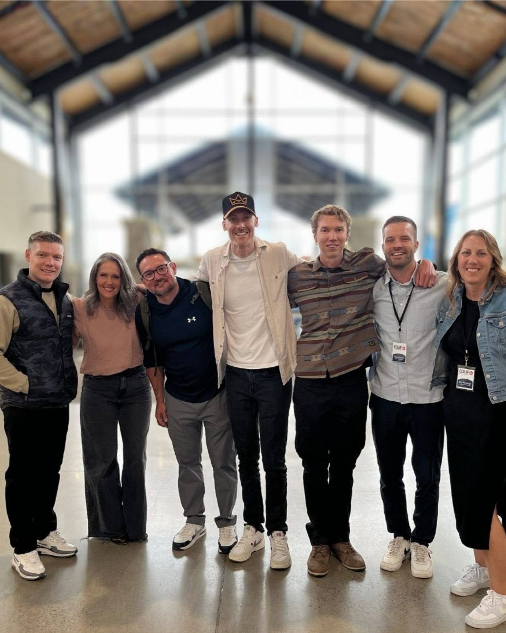 Group of seven smiling young adults standing closely together indoors in front of large windows, with some placing arms around each other's shoulders.