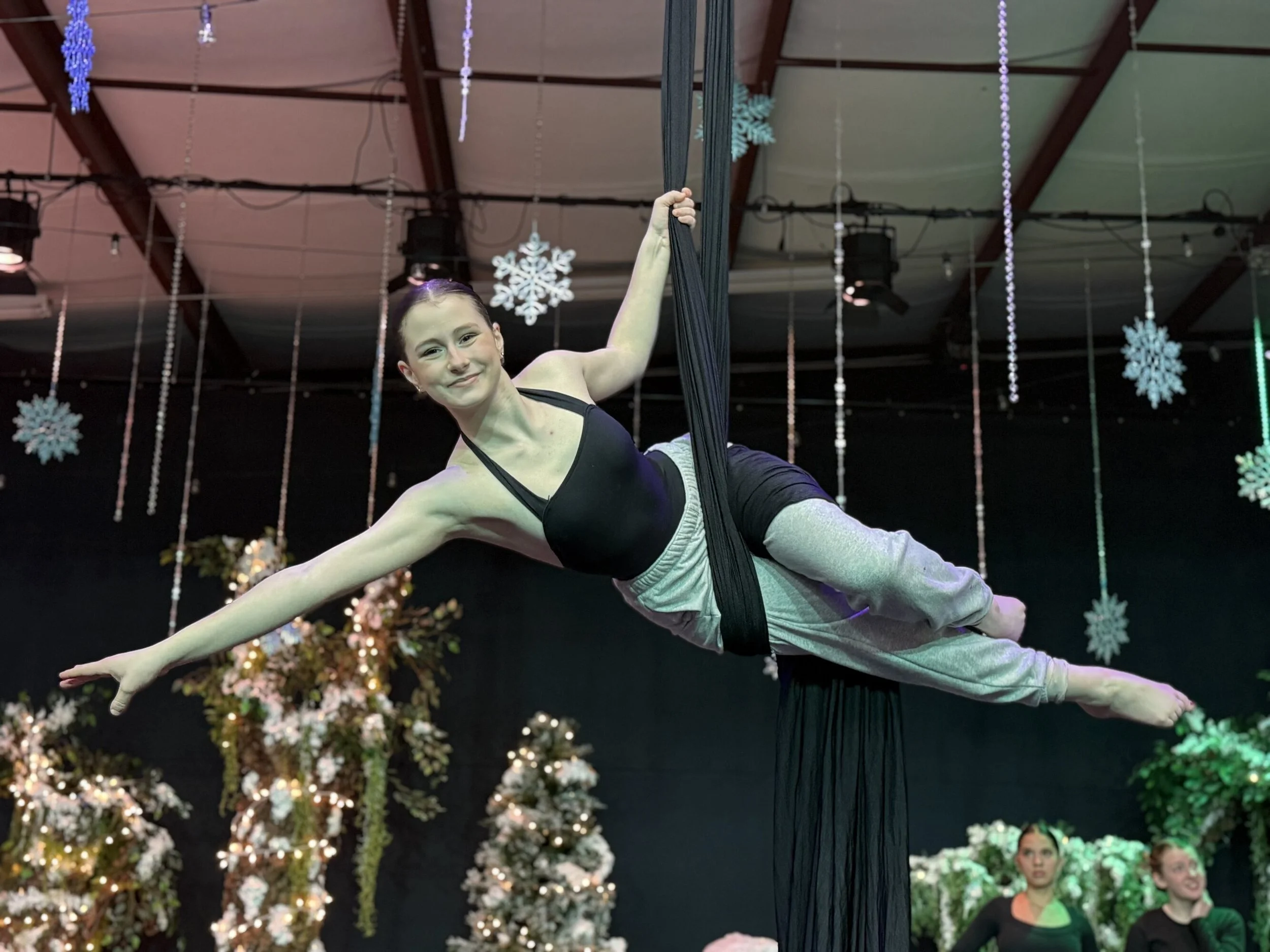 Aerial silk performer smiling while hanging horizontally on black silks decorated with Christmas trees and snowflake ornaments in the background.
