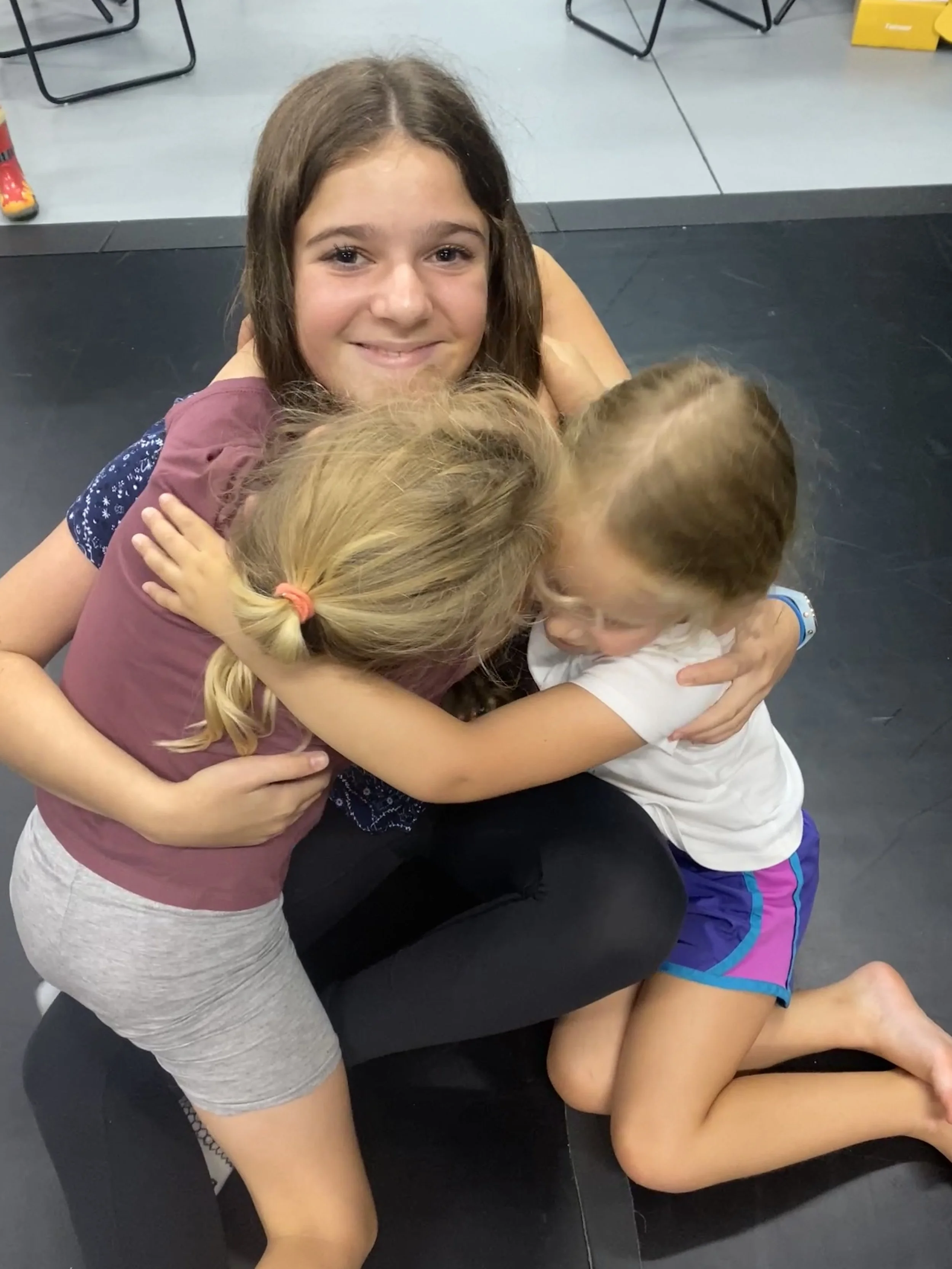 A young woman sitting on a black mat hugging three young girls who are playfully hugging her.