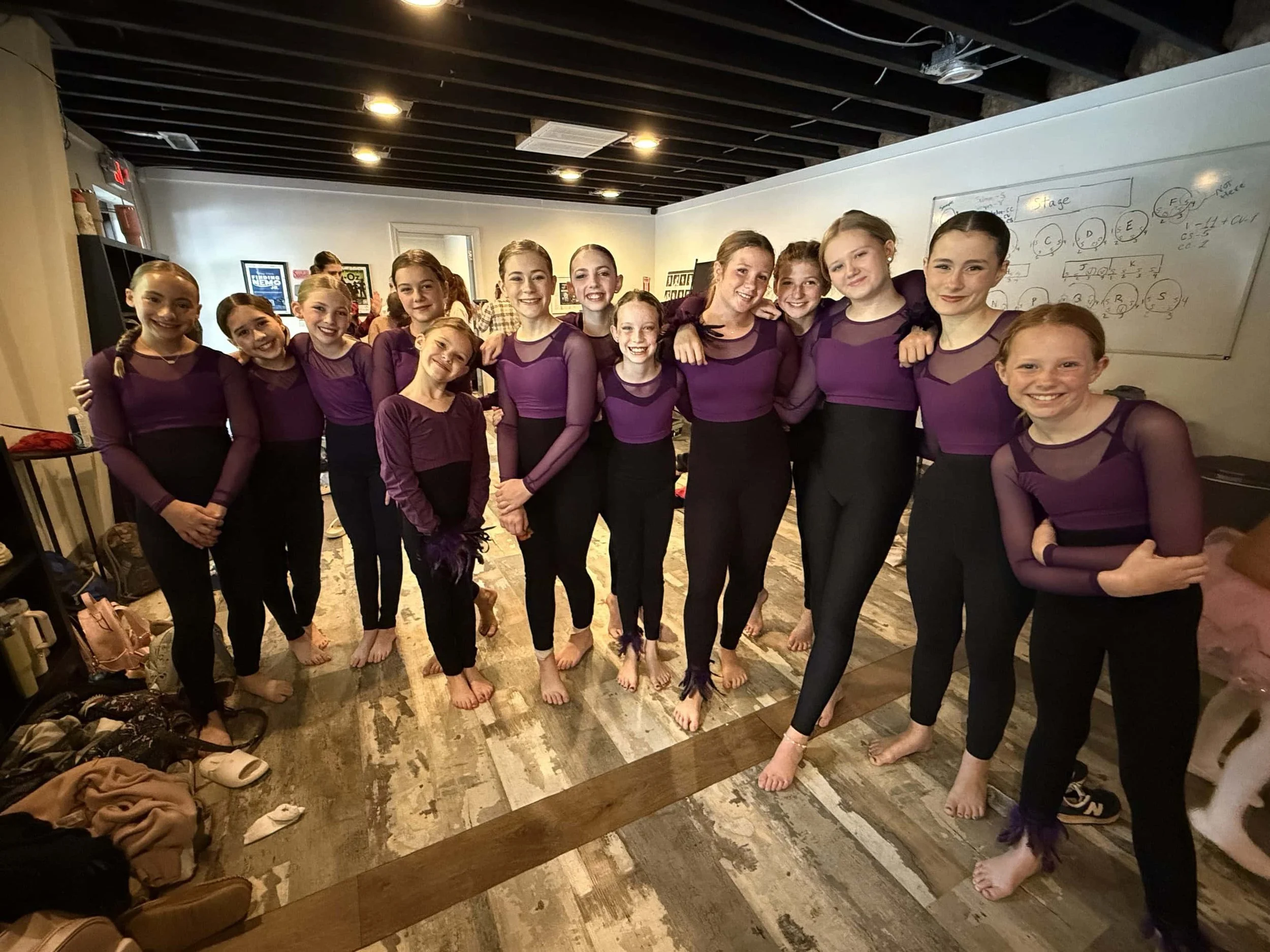 Group of young girls in matching purple and black dance costumes standing together in a dance studio, smiling and posing for a photo.