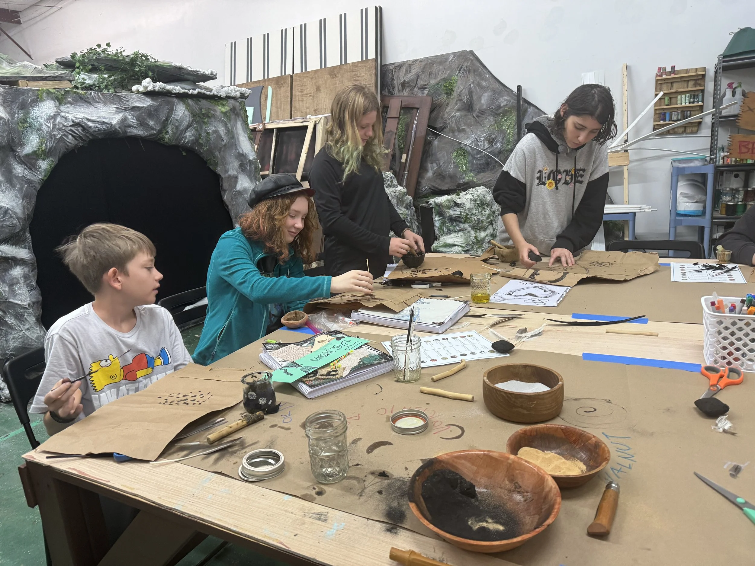 Children working on an art project at a workshop table with art supplies, papers, and carving tools.