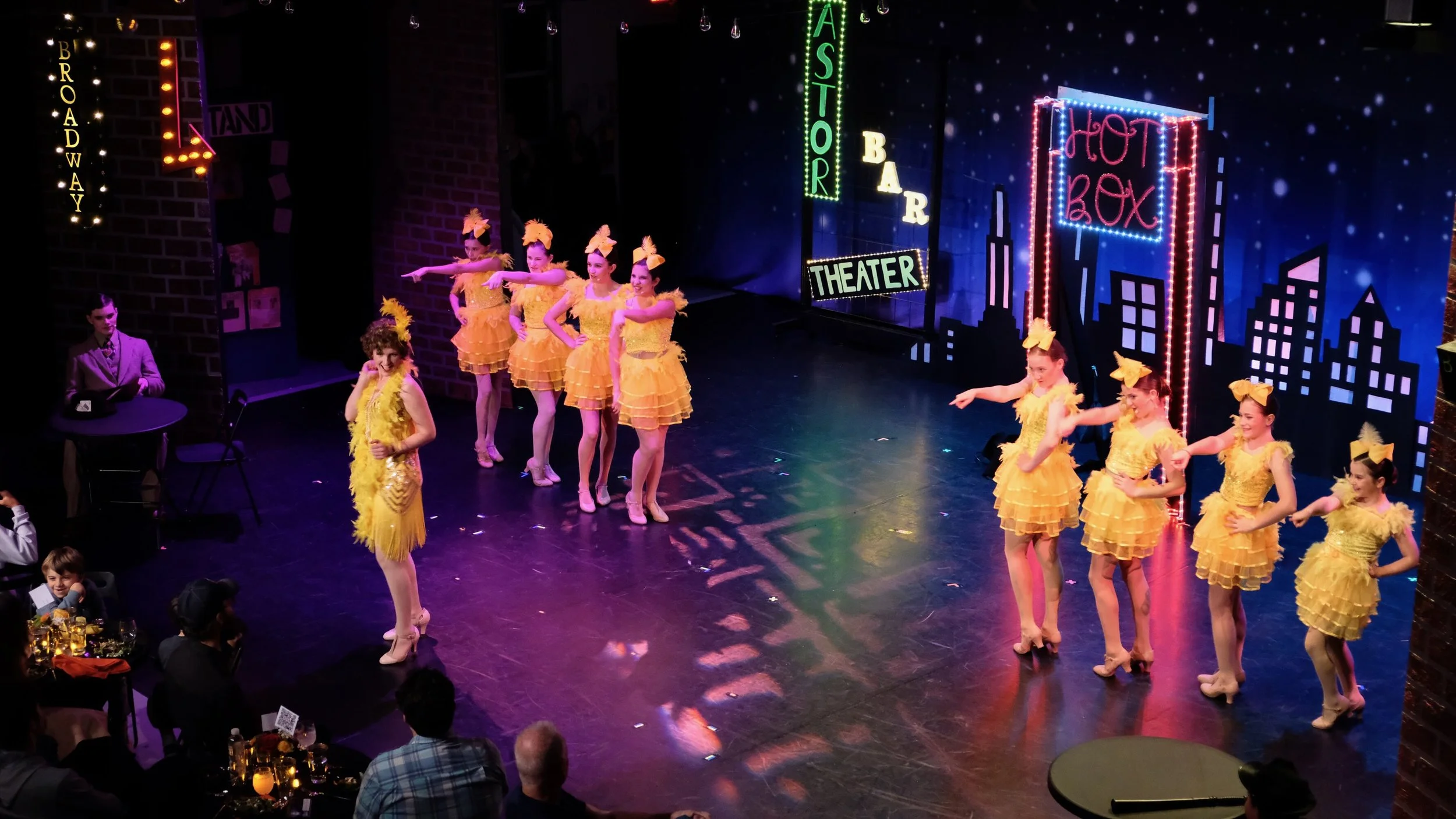 A stage performance with nine young girls dressed in yellow costumes dancing. The background features cityscape neon signs, including 'Hot Box' and 'Theater', and Broadway and Astor signs. Audience members are watching from tables.