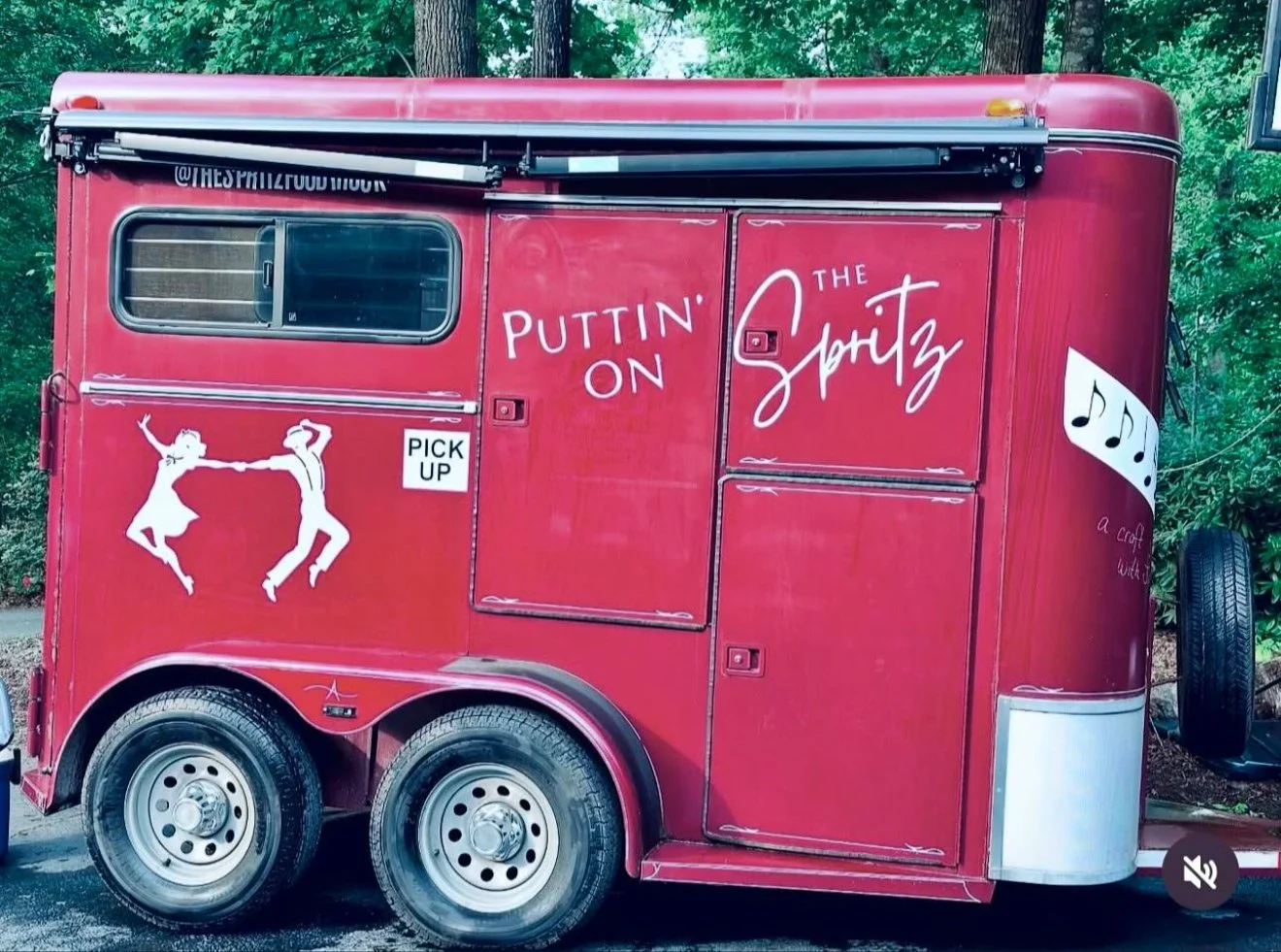 A red food trailer with white text and illustrations. It has a window, wheels, and a sign saying ''PICK UP''. The text on the trailer reads ''PUTTIN' ON THE SPOTZ'' and ''a craft with''. There is a graphic of two dancing figures holding hands and a m