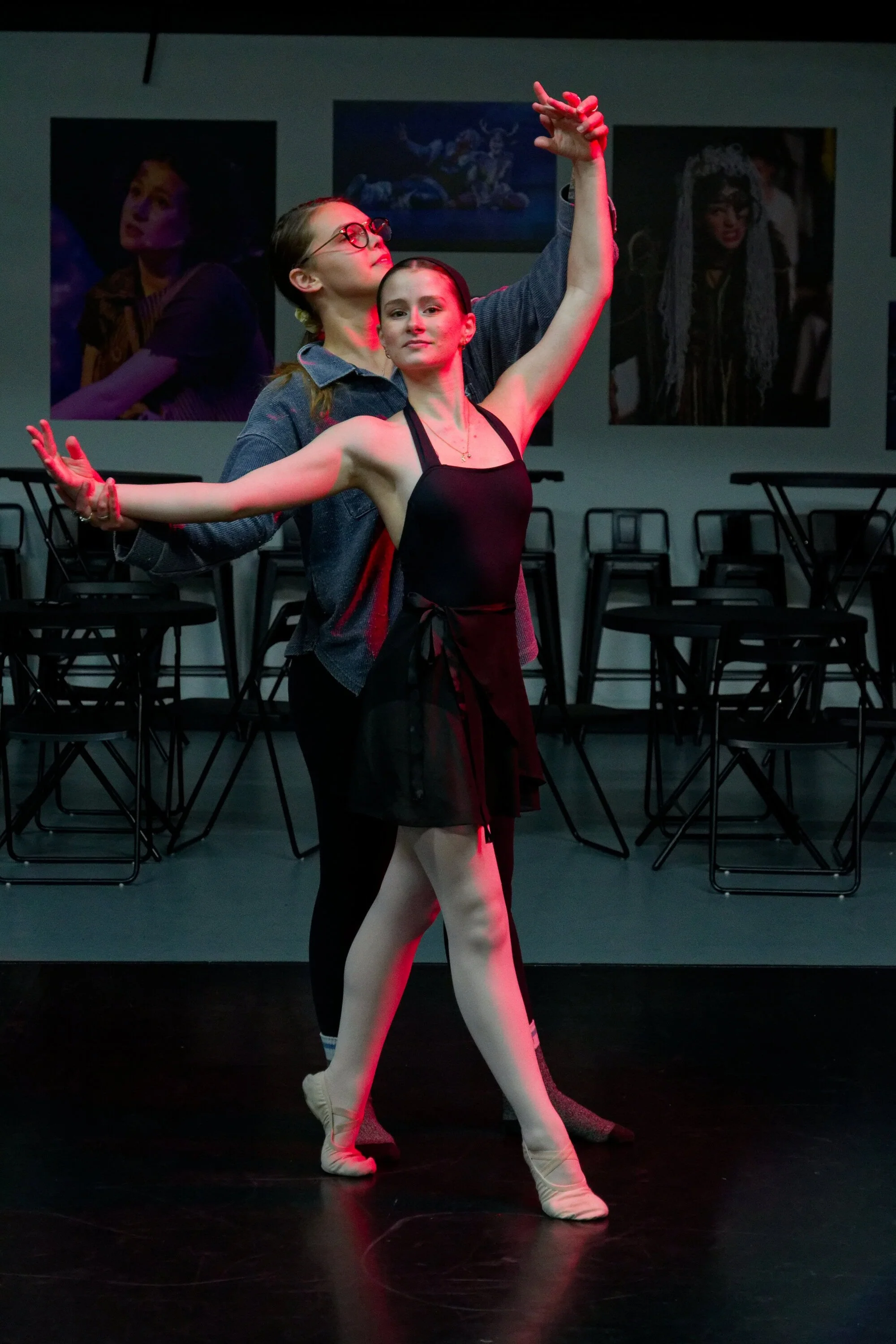 Two ballet dancers practicing on stage, with one standing en pointe and the other behind, both in dance poses, in a studio with chairs and framed photographs or artwork on the wall.