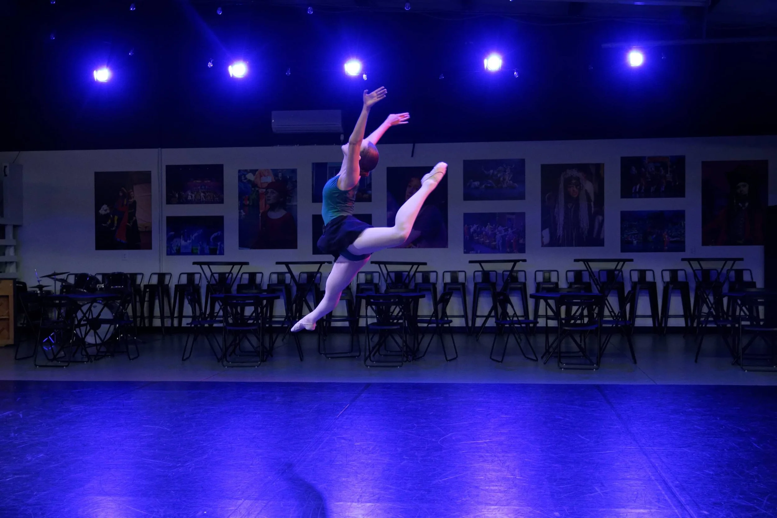 A ballet dancer mid-air in a dance studio with purple lighting, black chairs lined along the back wall, and framed pictures hanging on the wall.