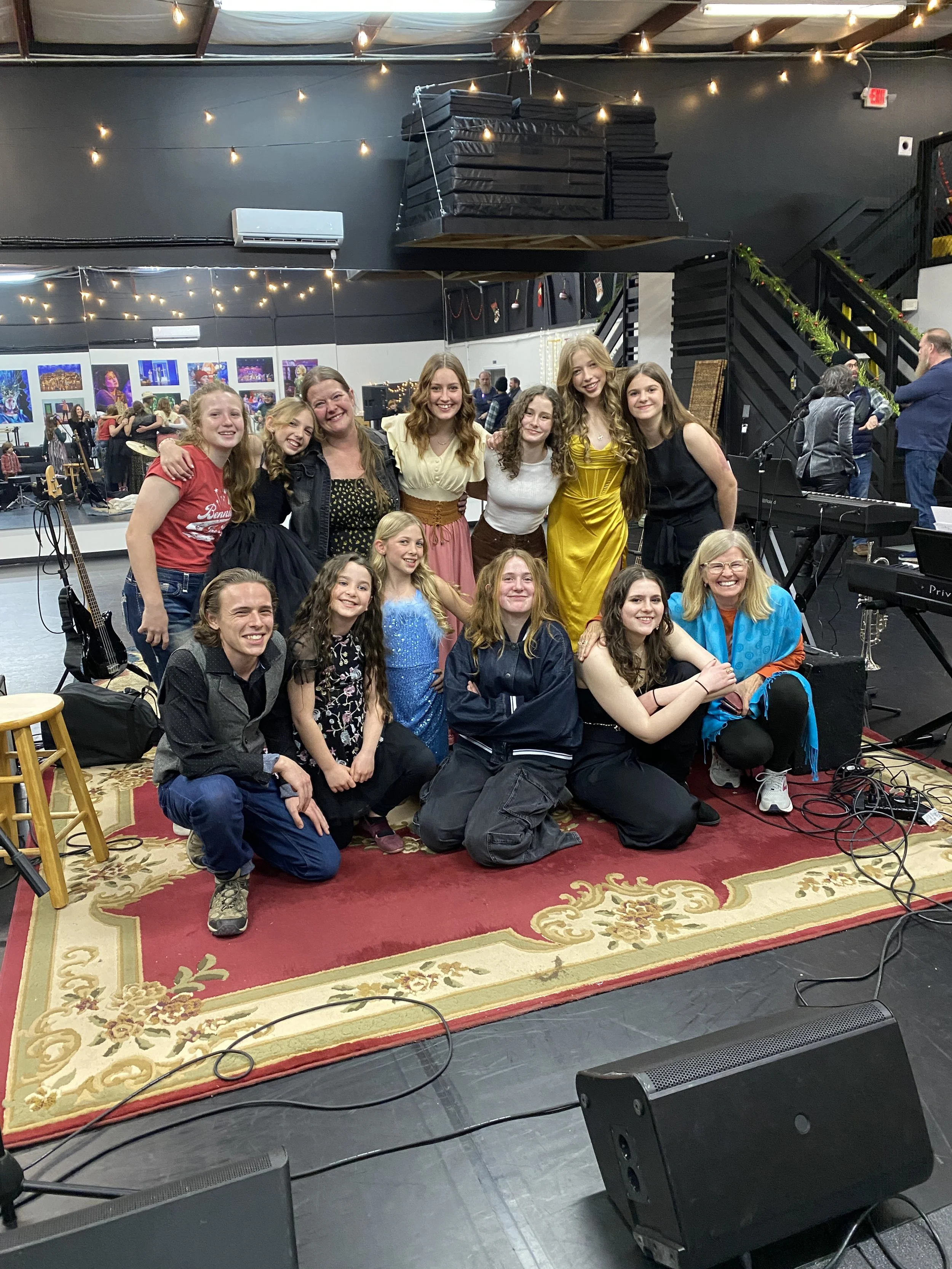 A group of children and a woman are gathered together on a red patterned rug inside a music venue, smiling for a photo. In the background, there are musical instruments, microphones, and stage lighting, with people socializing and performing.
