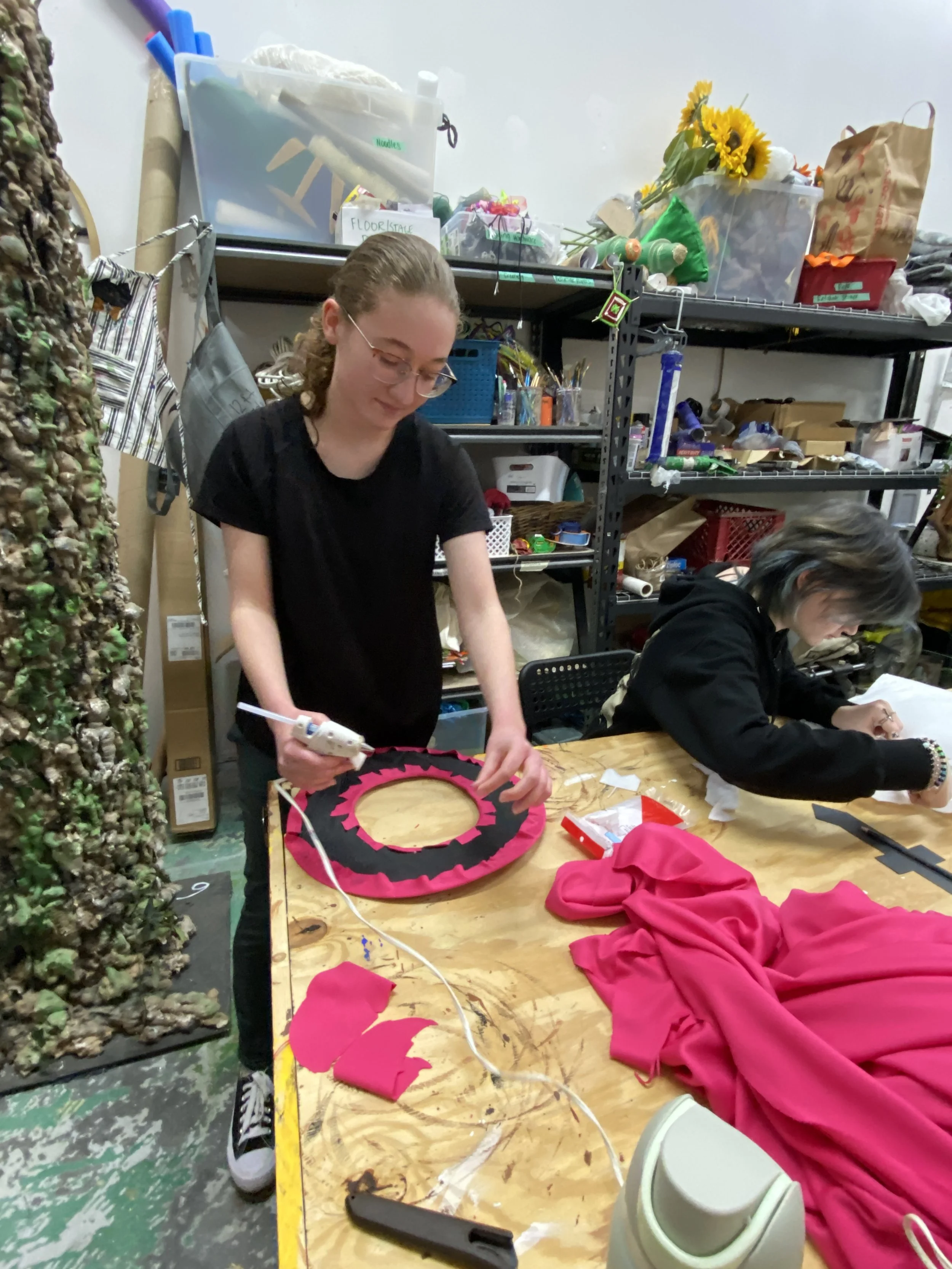 Two women working on craft projects at a workshop table, surrounded by sewing supplies, fabric, and shelves filled with various items.