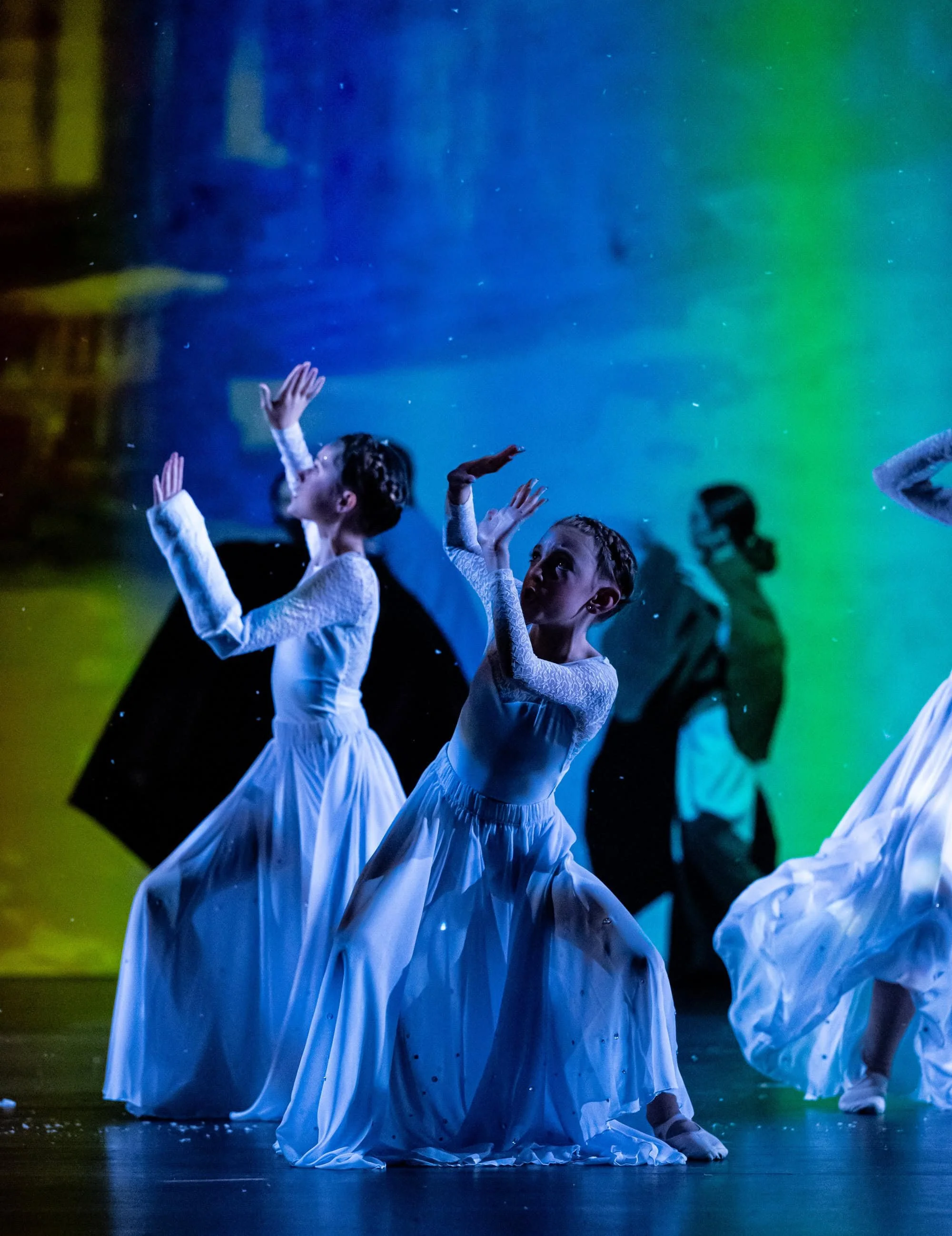 Children in white dance costumes performing a dance routine on stage with colorful background lighting.