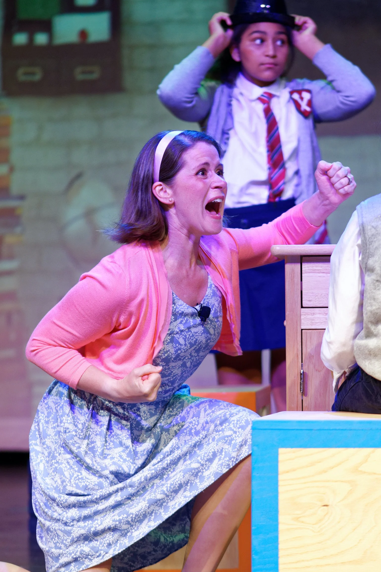 A woman wearing a pink cardigan and a floral dress, appearing angry and yelling, with her fist clenched, in a theatrical performance. Behind her, a young girl dressed as a student with a striped tie and a cardigan, is adjusting a black hat.