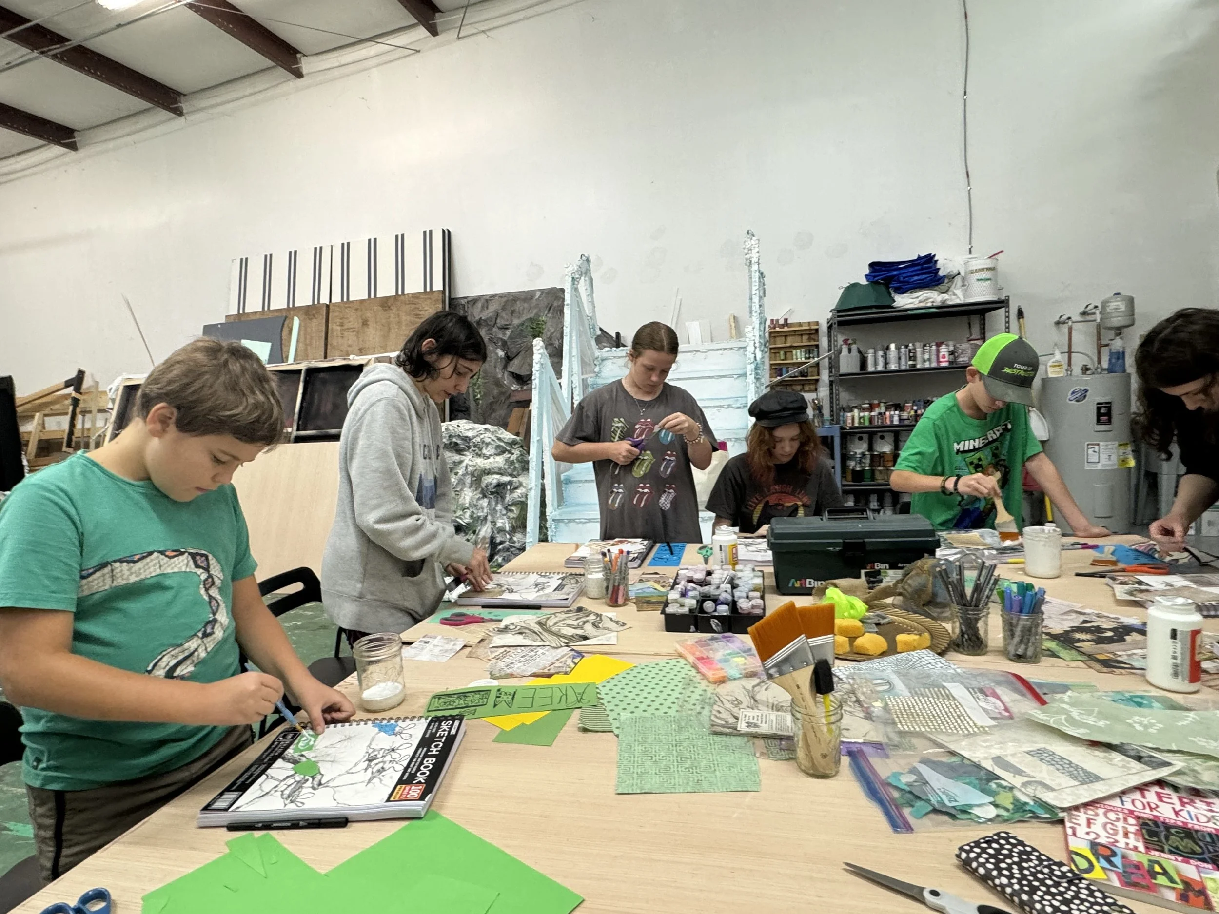Children and teenagers engaging in arts and crafts activities at a large table in an art studio or classroom, with various art supplies and materials on the table.
