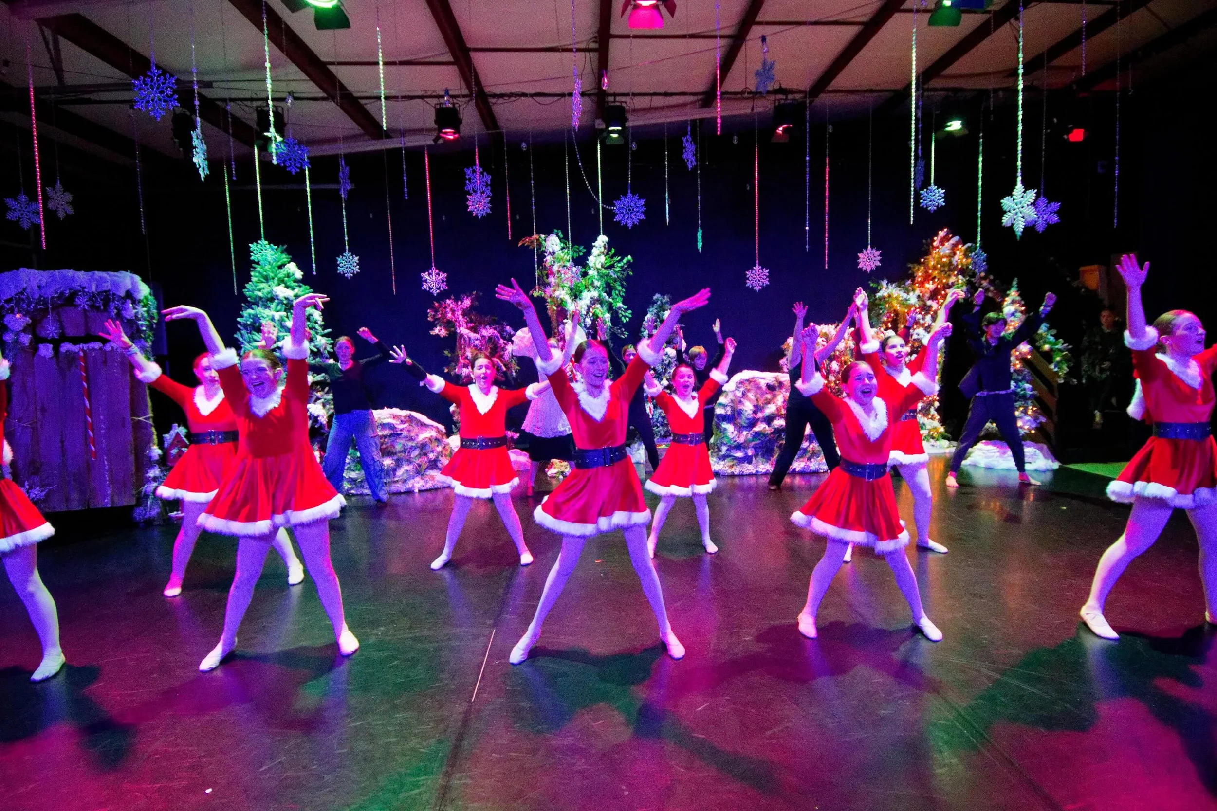 Children dressed as Santa Clauses performing a dance on stage decorated with Christmas trees, snowflakes, and colorful lights.