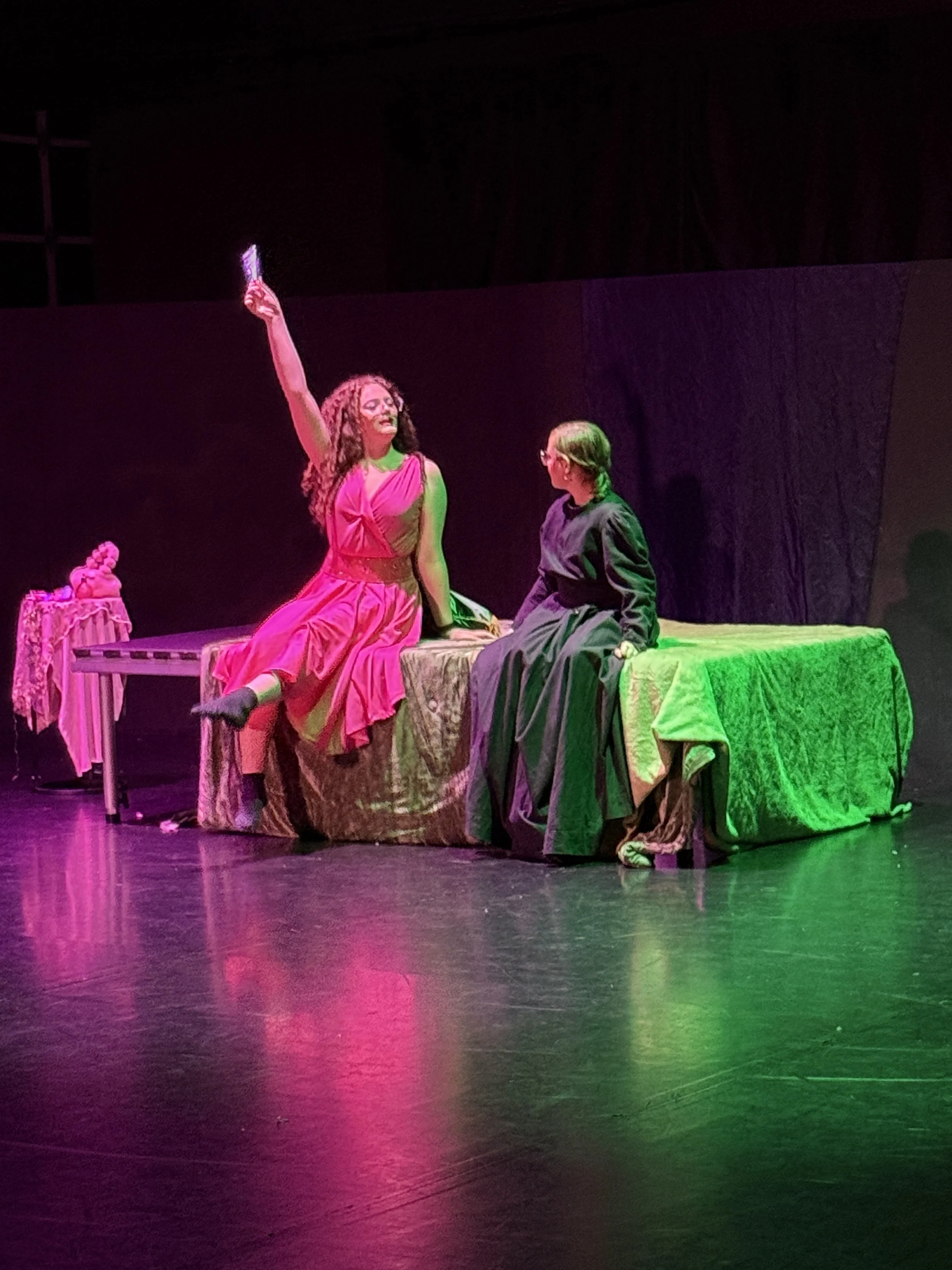 Two women sit on a bed in a theatrical stage setting, with one woman raising a fan and wearing a pink dress, and the other woman wearing a black dress, engaging in conversation. The scene is lit with pink and green lighting.
