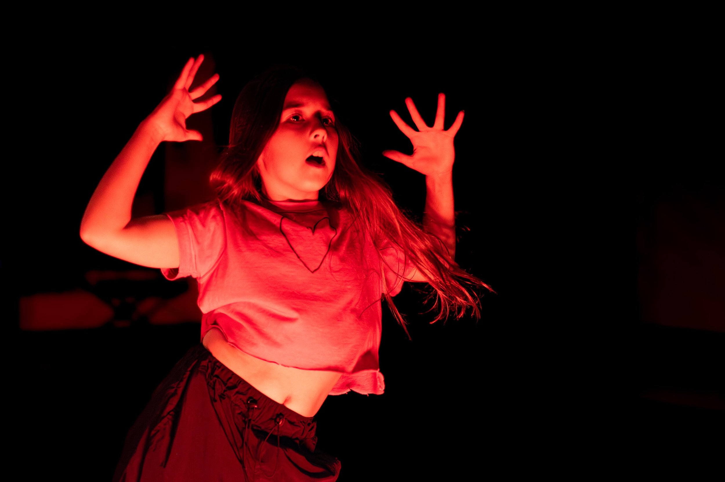Young girl with long hair raising her hands with a surprised or scared expression, illuminated by red lighting against a dark background.