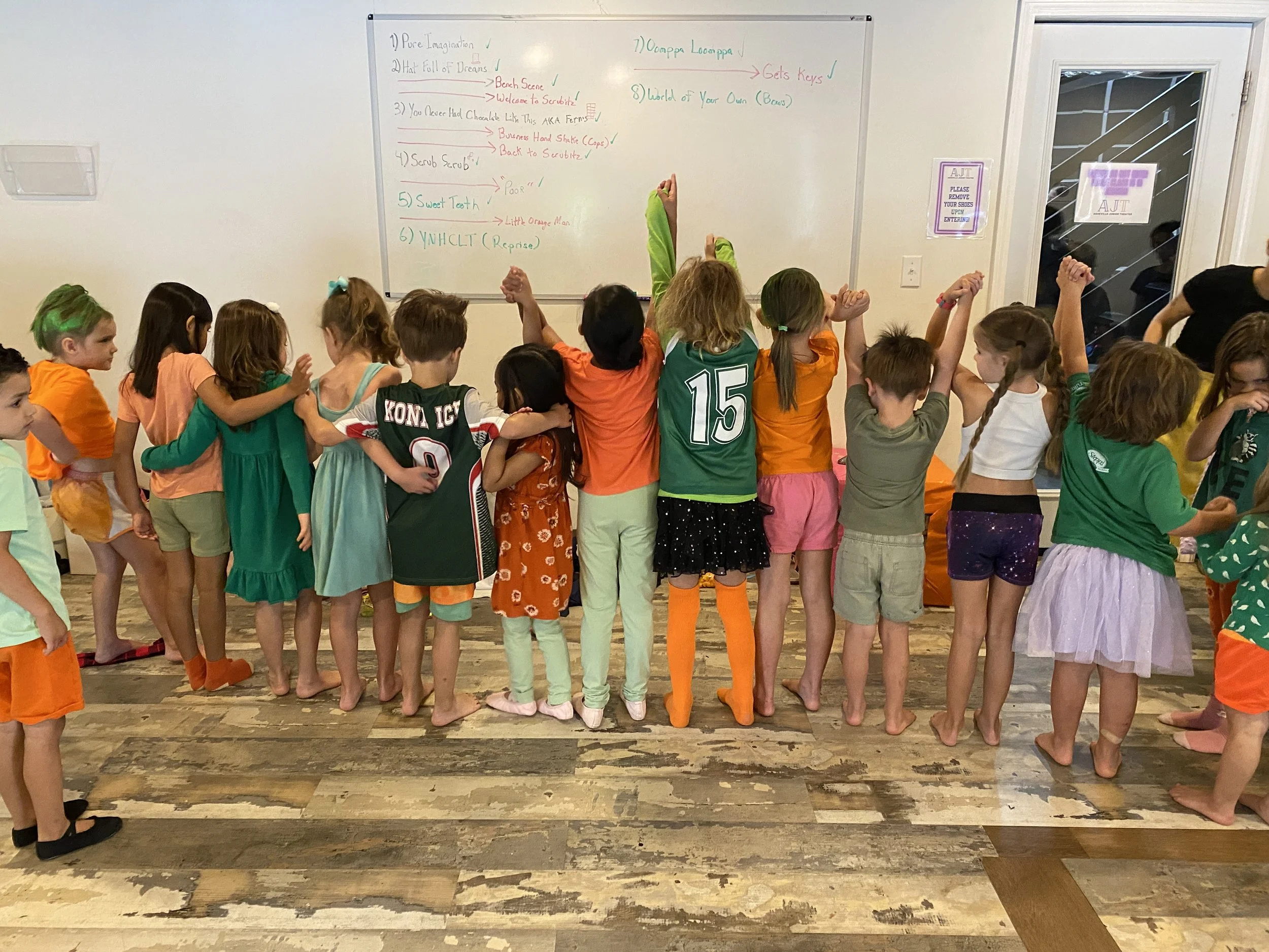 A group of children with diverse hair colors and styles standing together with their backs to the camera, some with arms raised, in front of a whiteboard with colorful writings.