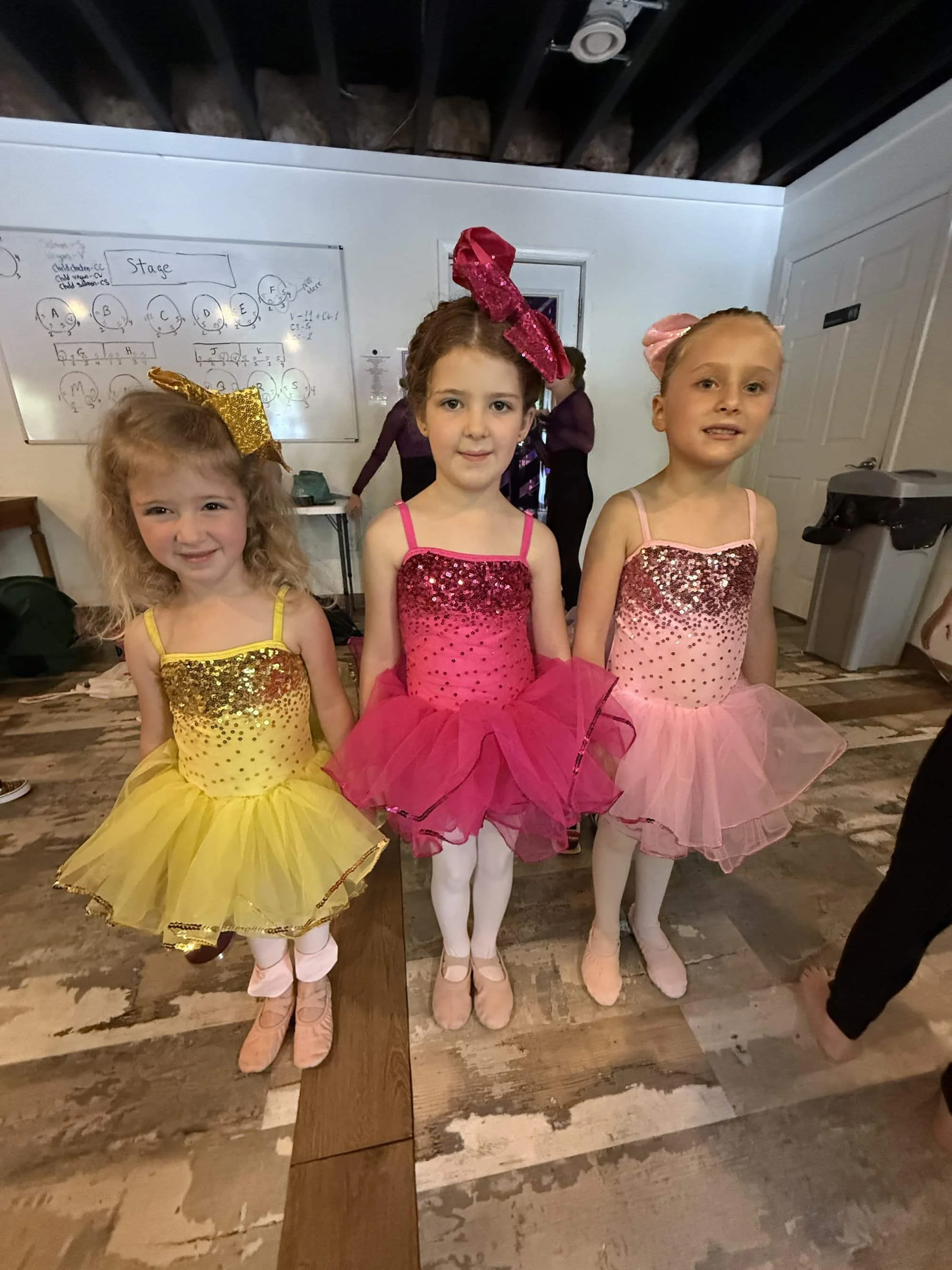 Three young girls dressed in colorful tutus and leotards, standing together indoors, ready for a dance performance or recital.