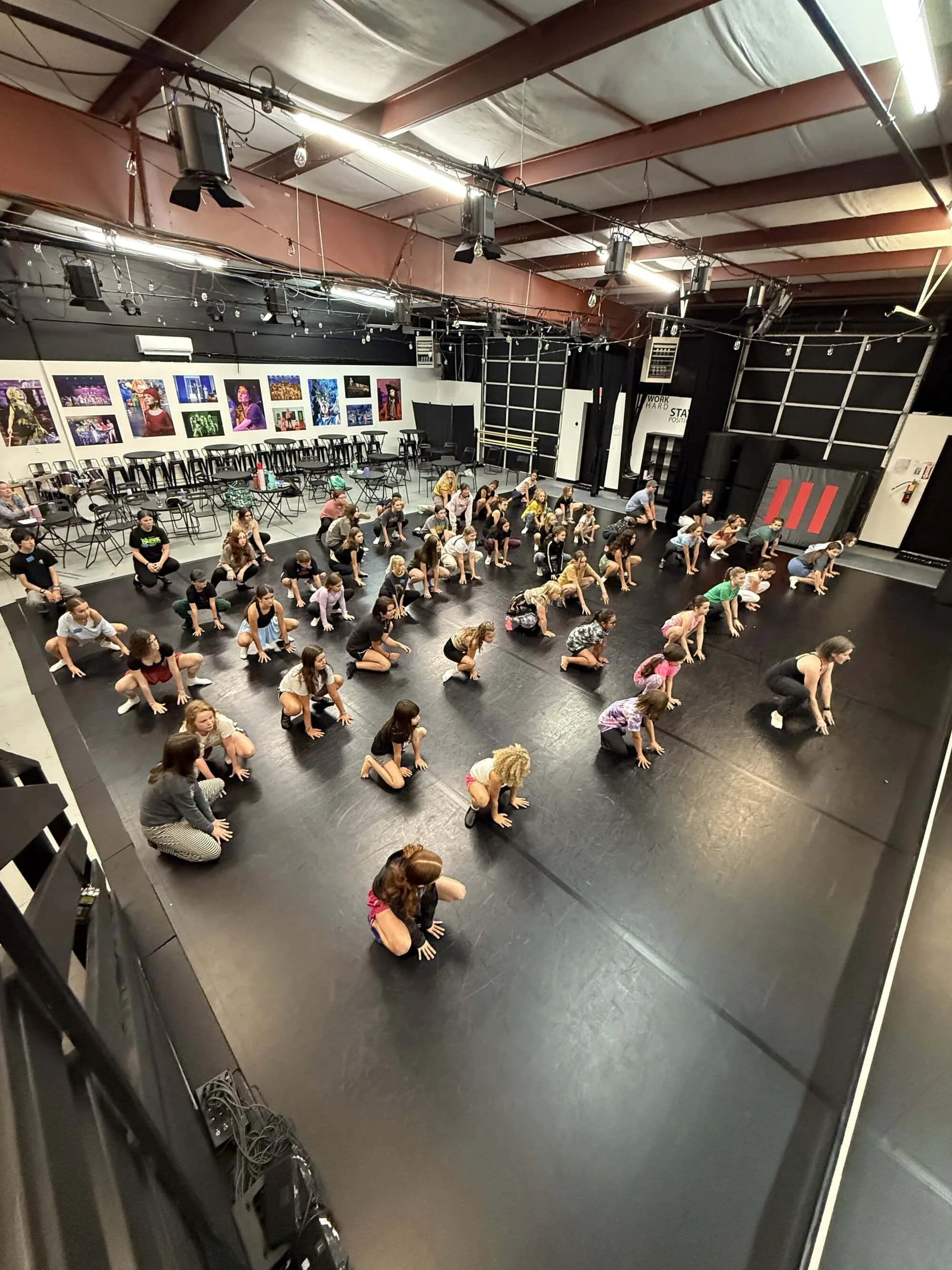 Group of children and adults practicing dance moves in a black dance studio, with posters on the wall and a row of empty chairs and tables along the back wall.