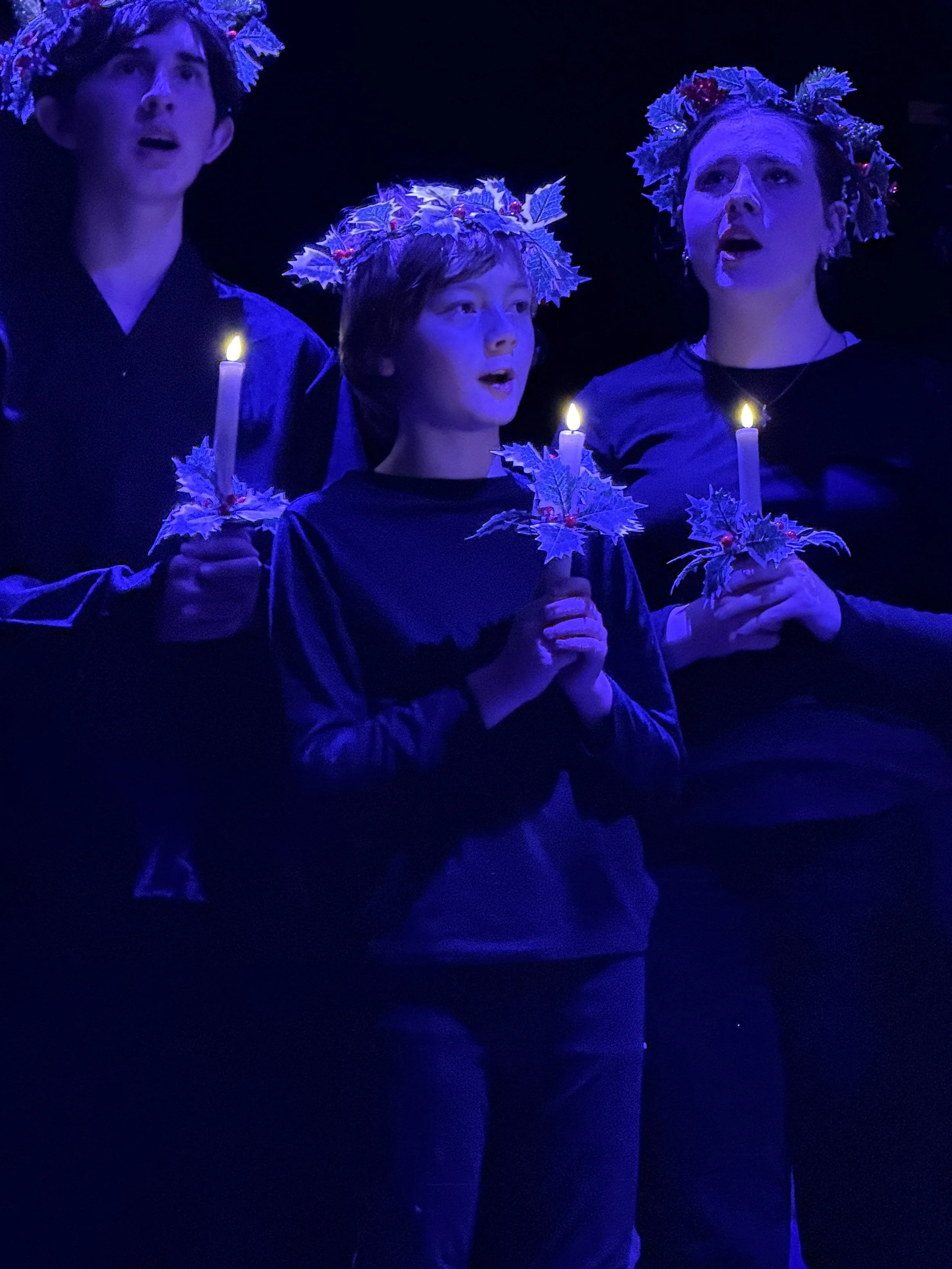 Three people singing with candles and holly leaf headpieces during a performance, illuminated by blue stage lighting.