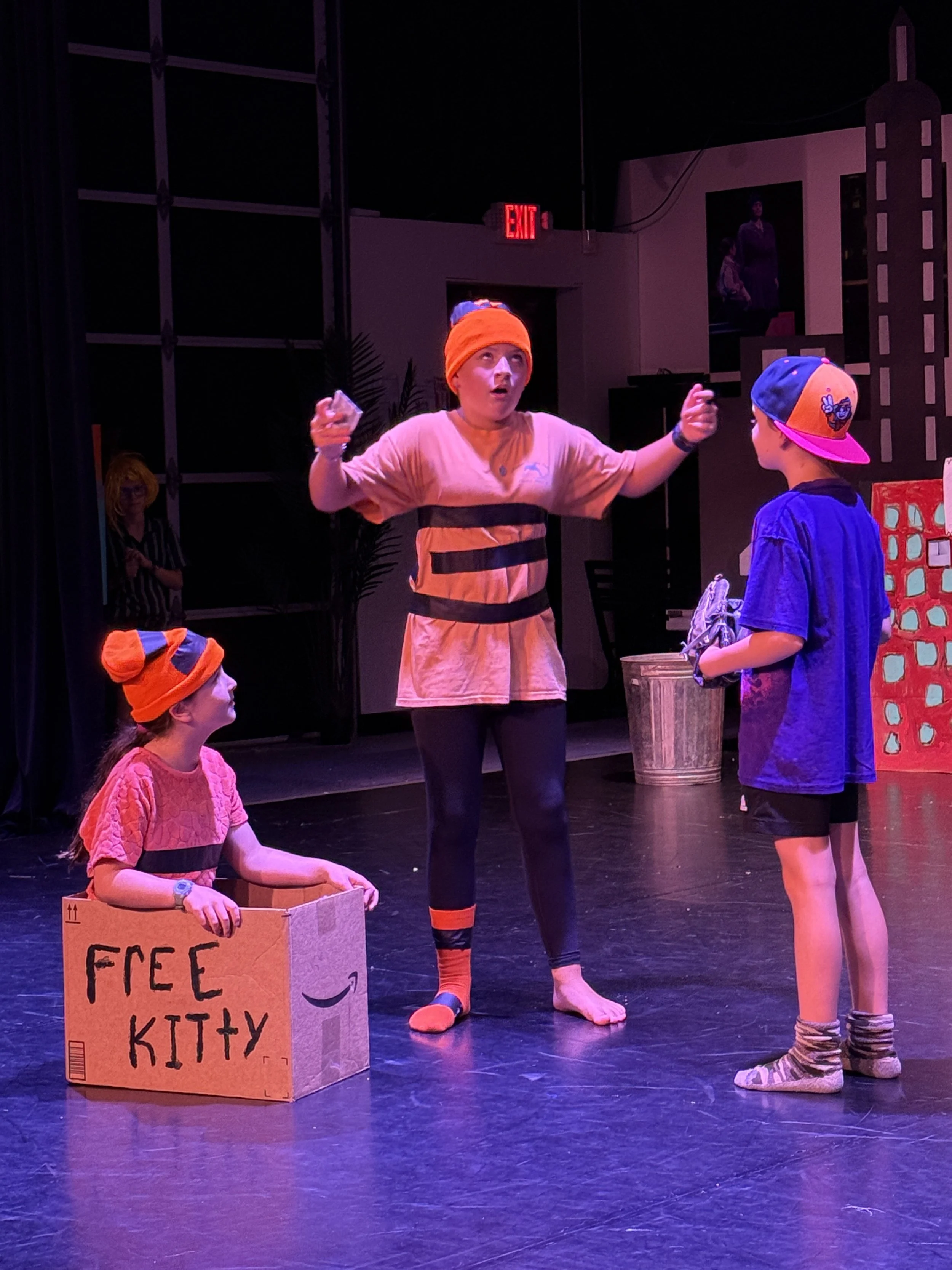 Three children and an adult on stage, with two children wearing orange hats and a cardboard box labeled 'FREE KITTY'. The adult is gesturing while speaking, and the children are listening. The scene appears to be from a theatrical performance.