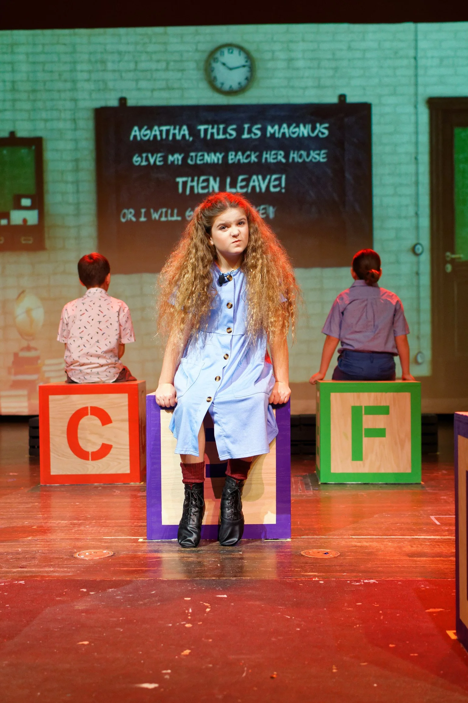 A young girl with long curly hair standing on a stage with colorful blocks, in front of a blackboard with a play script, while two children sit on blocks facing away. The scene appears to be part of a theatrical performance.